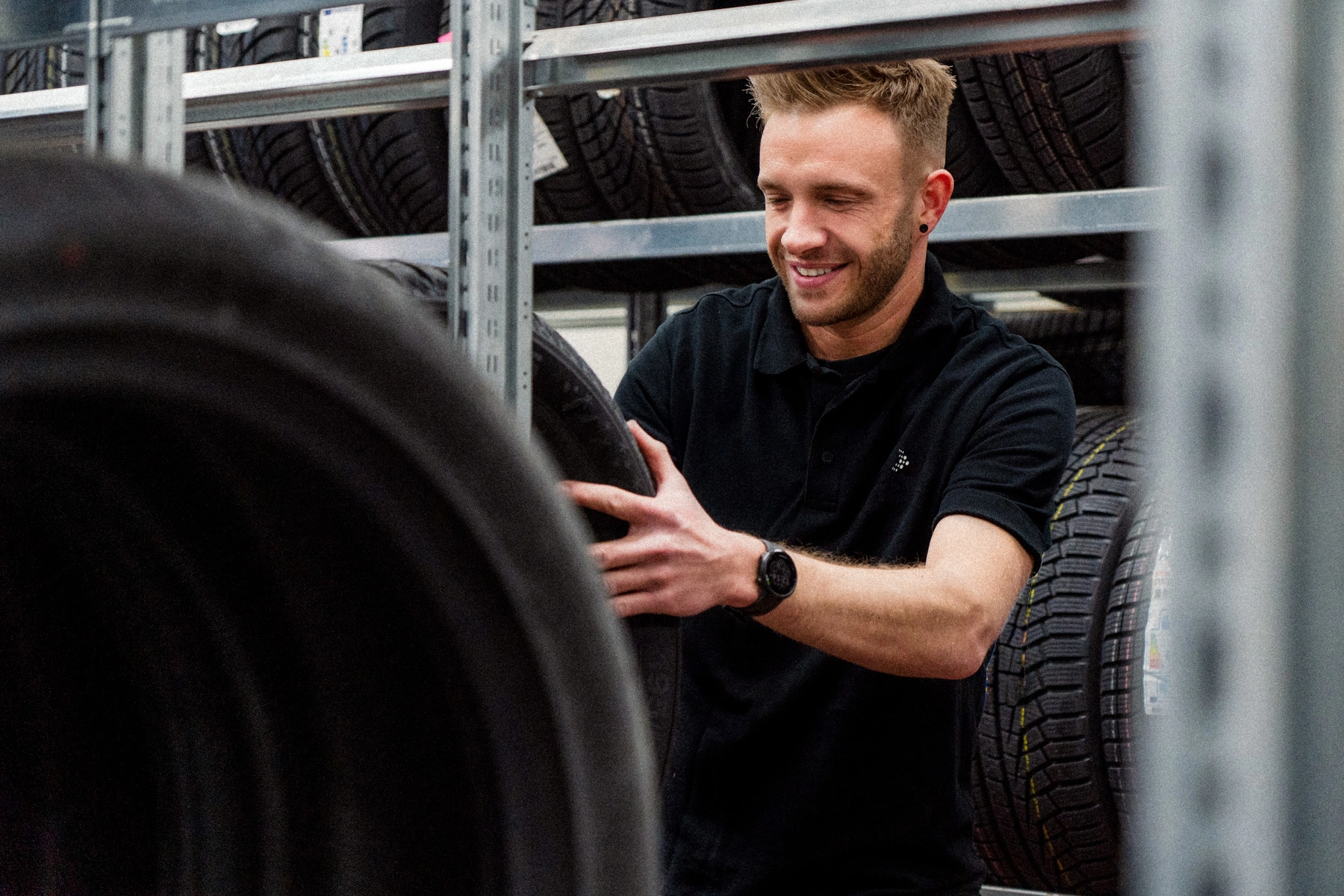 Eine Aufnahme im Reifenlager des Autohauses. Ein Teiledienst-Mitarbeiter in schwarzen AVEMO Polo-Shirt lädt einen Reifen in das Regal ein.