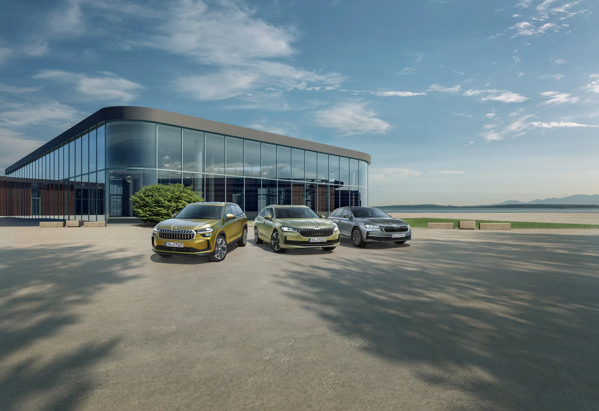 Drei Škoda-Modelle – ein goldener Kodiaq SUV, ein grüner Superb Combi und ein silberner Octavia Combi – stehen auf einem sonnigen Vorplatz vor einem modernen Glasgebäude am See. Der blaue Himmel und die Berge im Hintergrund schaffen eine offene Atmosphäre.