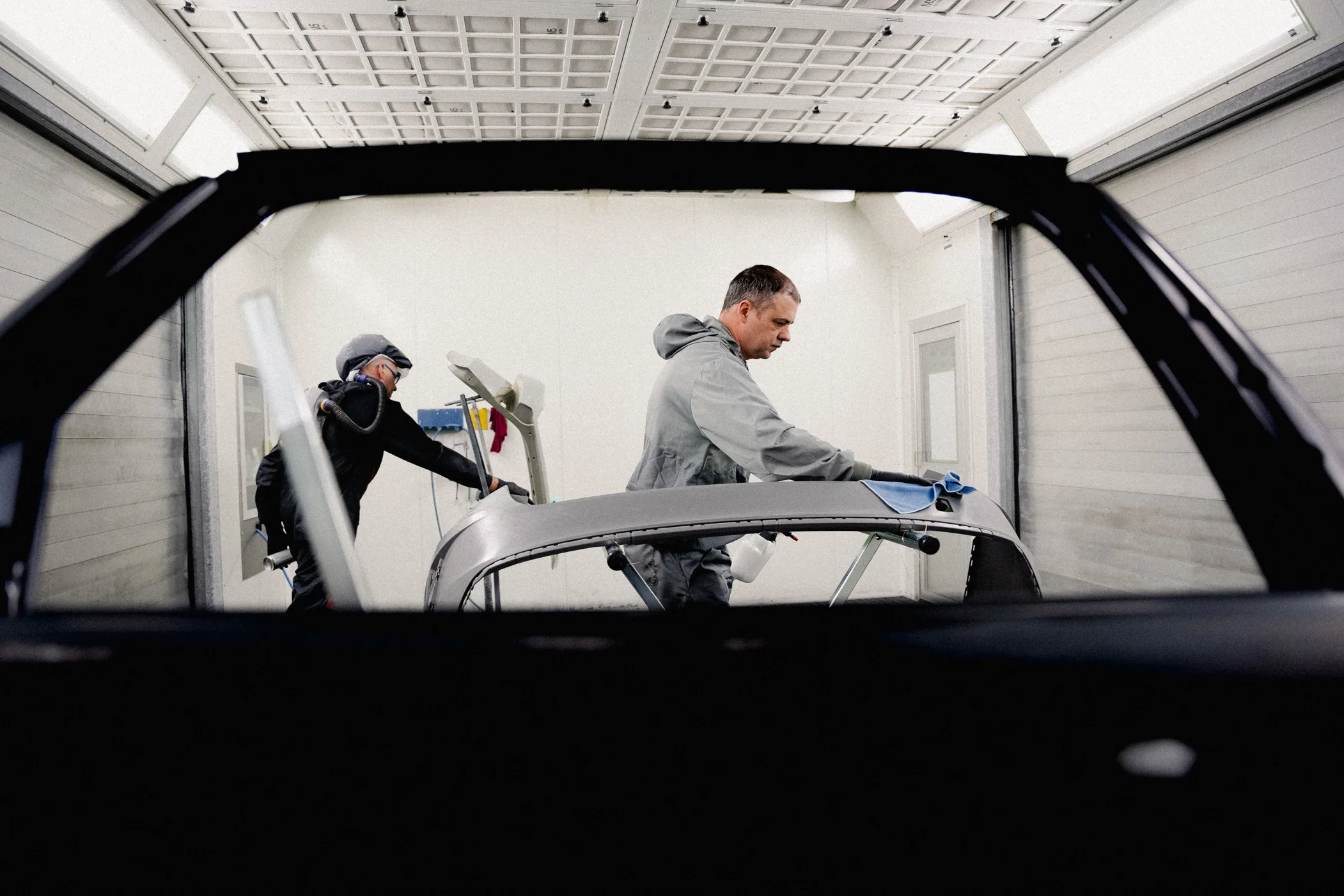 Zu sehen sind zwei Mitarbeiter in Schutzkleidung in einer Lackierkabine. Beide treffen Vorbereitungen an Karosserie-Bauteilen, um sie anschließend lackieren zu können. Das Bild wurde durch den Rahmen einer ausgebauten Tür hindruch aufgenommen.