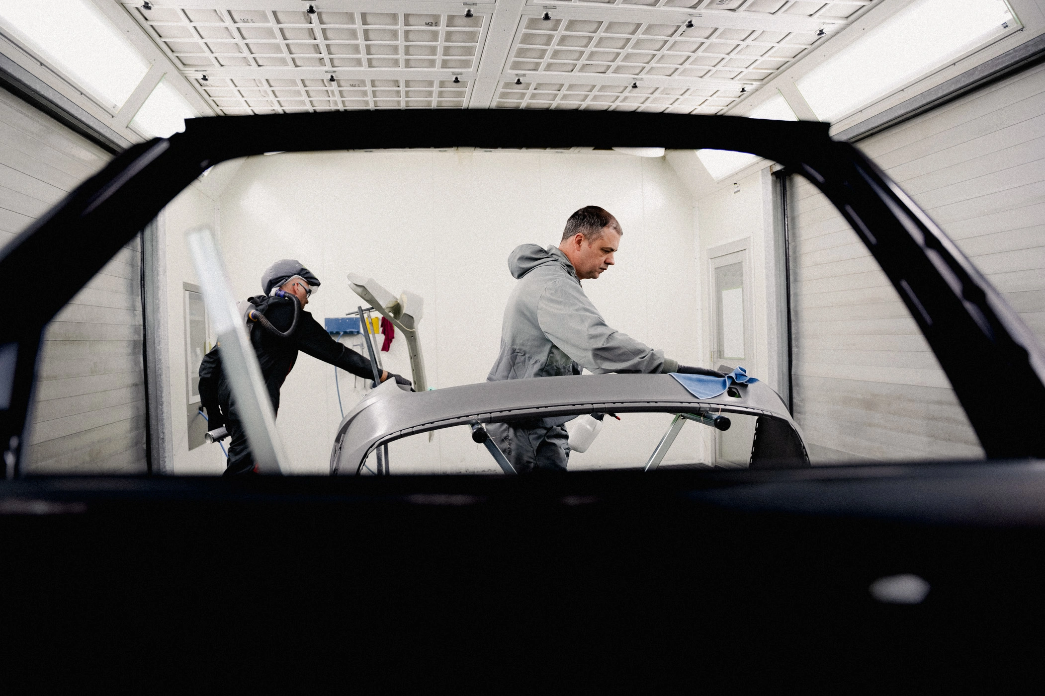 Lackierkabine m,m Zu sehen sind zwei Mitarbeiter in Schutzkleidung in einer Lackierkabine. Beide treffen Vorbereitungen an Karosserie-Bauteilen, um sie anschließend lackieren zu können. Das Bild wurde durch den Rahmen einer ausgebauten Tür hindruch aufgenommen.