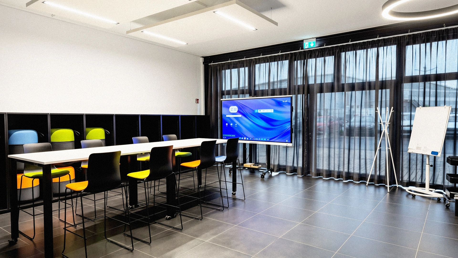 Ein moderner Besprechungsraum mit dunkelgrauem Fliesenboden. Zentral steht ein langer Stehtisch mit schwarz-bunten Barhockern. Links ein schwarzes Regal mit farbigen Sitzpoufs, rechts vor einer Fensterfront ein digitaler Monitor und ein Flipchart.