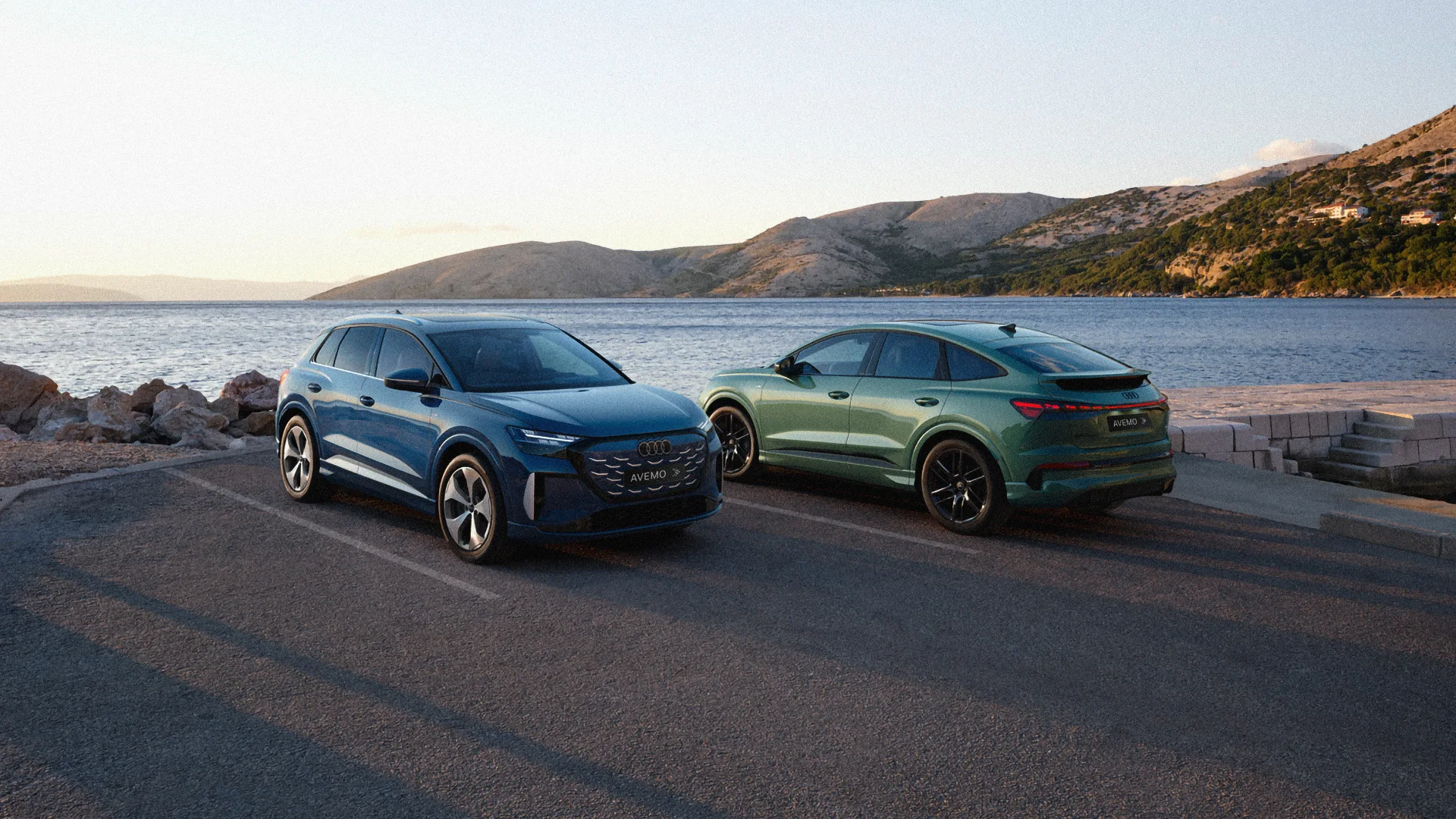Zwei Audi Q4 e-tron Modelle in Blau und Grün an einer Küstenstraße mit Meerblick bei Sonnenuntergang.