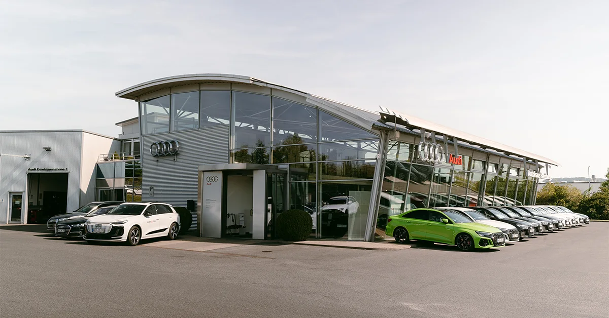Eine Aufnahme des Audi Autohauses in Haßfurt, die das Gebäude und Audi Fahrzeuge vor dem Gebäude sowie die Einfahrt zeigt.