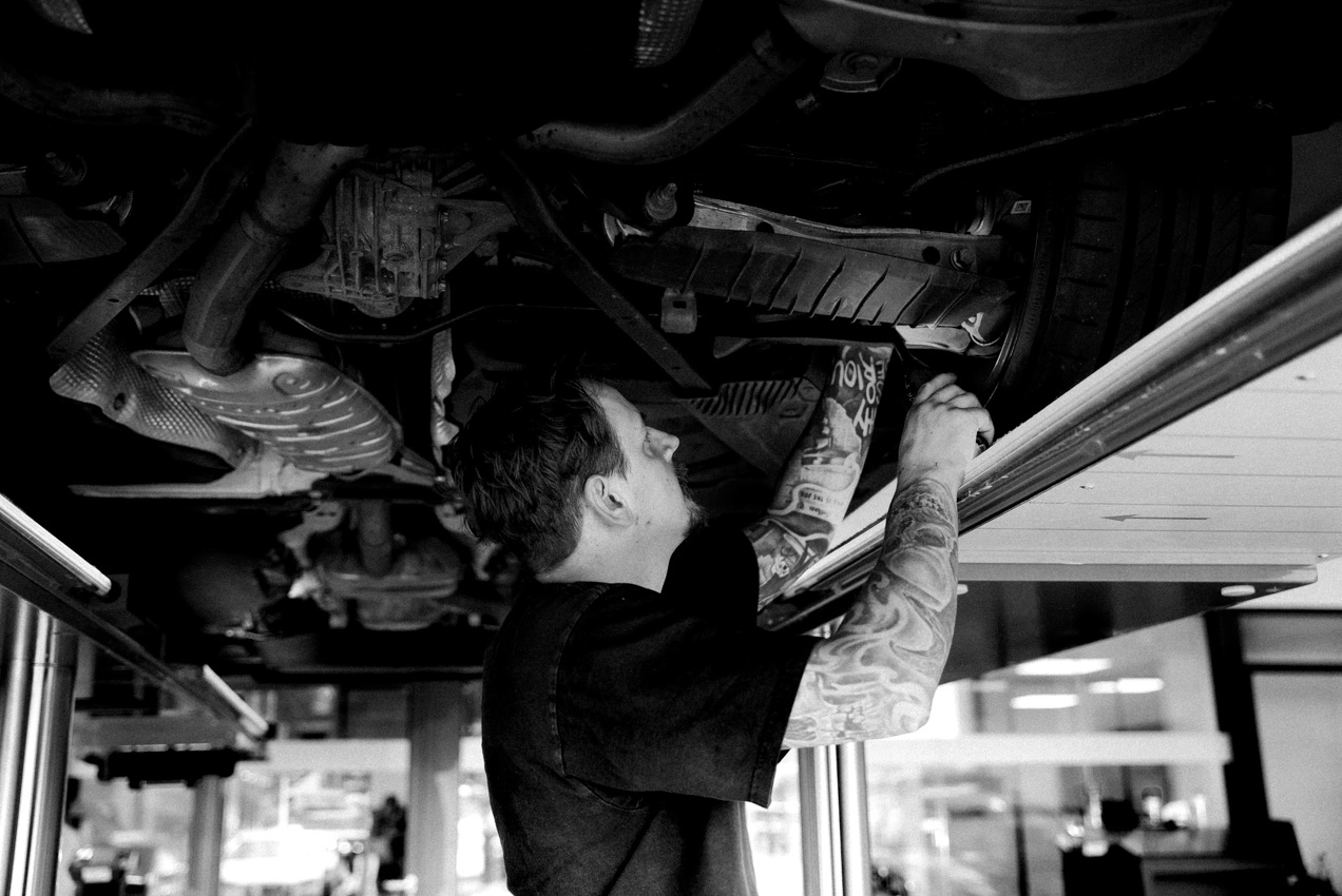 Man erkennt die Unterseite eines Fahrzeuges auf einer Hebebühne. Ein Werkstatt-Mitarbeiter steht unter dem Fahrzeug und inspiziert ein Bauteil an der Radaufhängung.