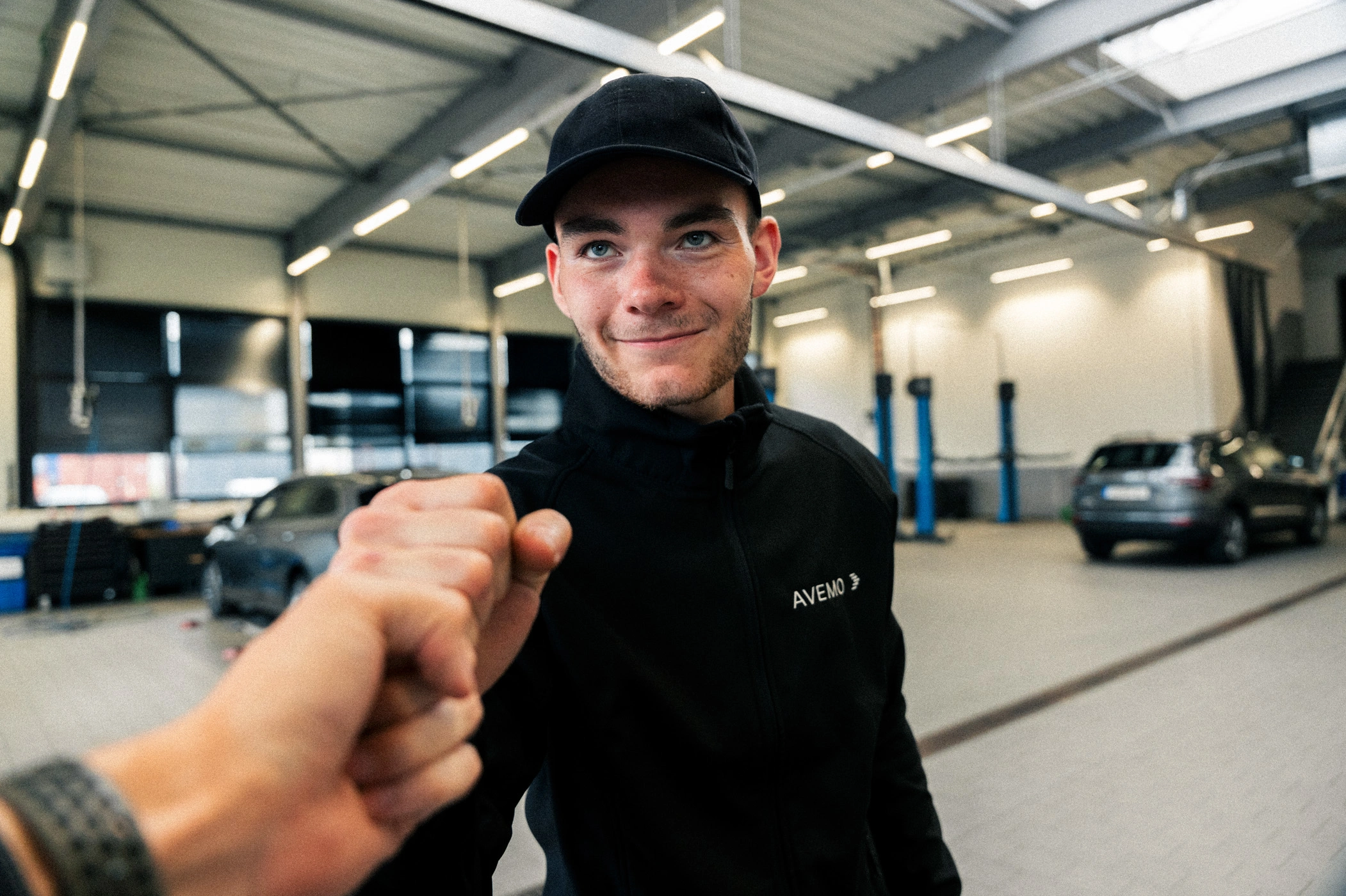 Werkstatt Fistbump m Eine Aufnahme eines Werkstatt-Mitarbeiters in schwarzer AVEMO Softshelljacke und mit Basecap, der mit dem Kameramann einen Fistbump durchführt.