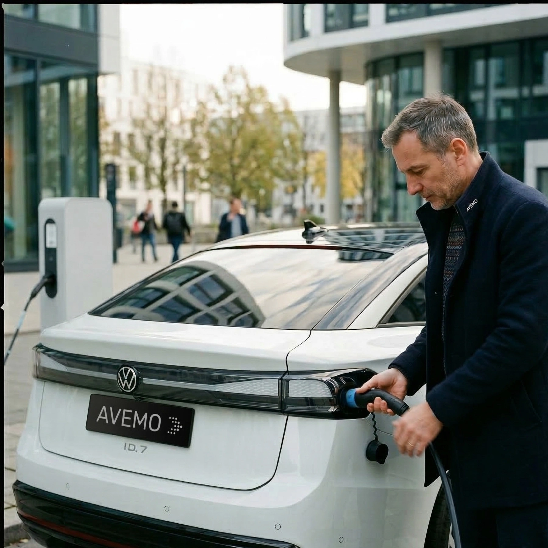 Ein Mann im dunklen Mantel steckt das Ladekabel in einen weißen Volkswagen ID.7 mit AVEMO-Kennzeichen. Das Elektroauto steht an einer Ladestation in einem modernen Stadtviertel mit Bürogebäuden im Hintergrund.