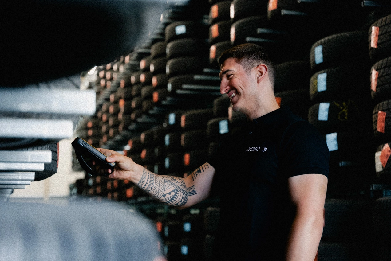 Teiledienst Reifenlager Scannen m Zu sehen ist ein Teiledienst-Mitarbeiter im Reifenlager. Er steht vor einem Regal mit Reifen und scannt mit dem Gerät in seiner Hand ein Label auf einem Reifen ein.