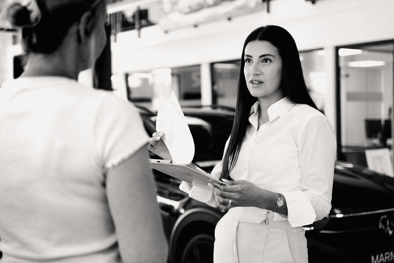 Information Konversation w,w-3 Eine Service-Mitarbeiterin und eine Kundin stehen zusammen im Showroom des Autohauses. Die Mitarbeiterin hält ein Klemmbrett mit Datenblättern in den Händen. Beide unterhalten sich miteinander. Im Hintergrund sieht man Büros und ein Neufahrzeug.