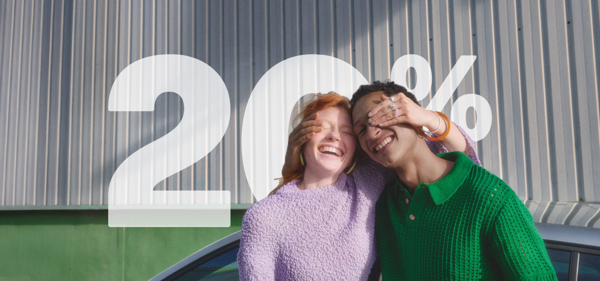 Ein lächelndes Paar steht vor einer grauen Blechwand. Eine Person mit dunklen Haaren bedeckt die Augen der rothaarigen Person, die laut lacht. Im Vordergrund überlagert eine große weiße Zahl "20%" die Szene und deutet auf einen Rabatt oder Vorteil hin.