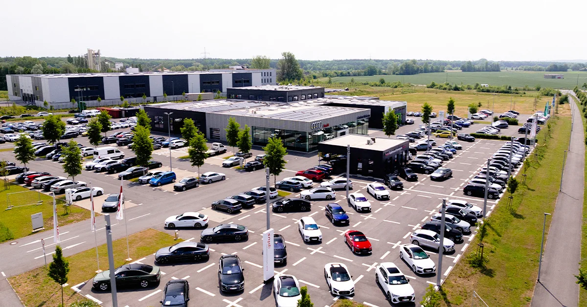 Eine Aufnahme des Audi Autohauses in Schweinfurt, die das Gebäude, den Parkplatz und Audi Fahrzeuge vor dem Gebäude, sowie die Einfahrt zeigt.