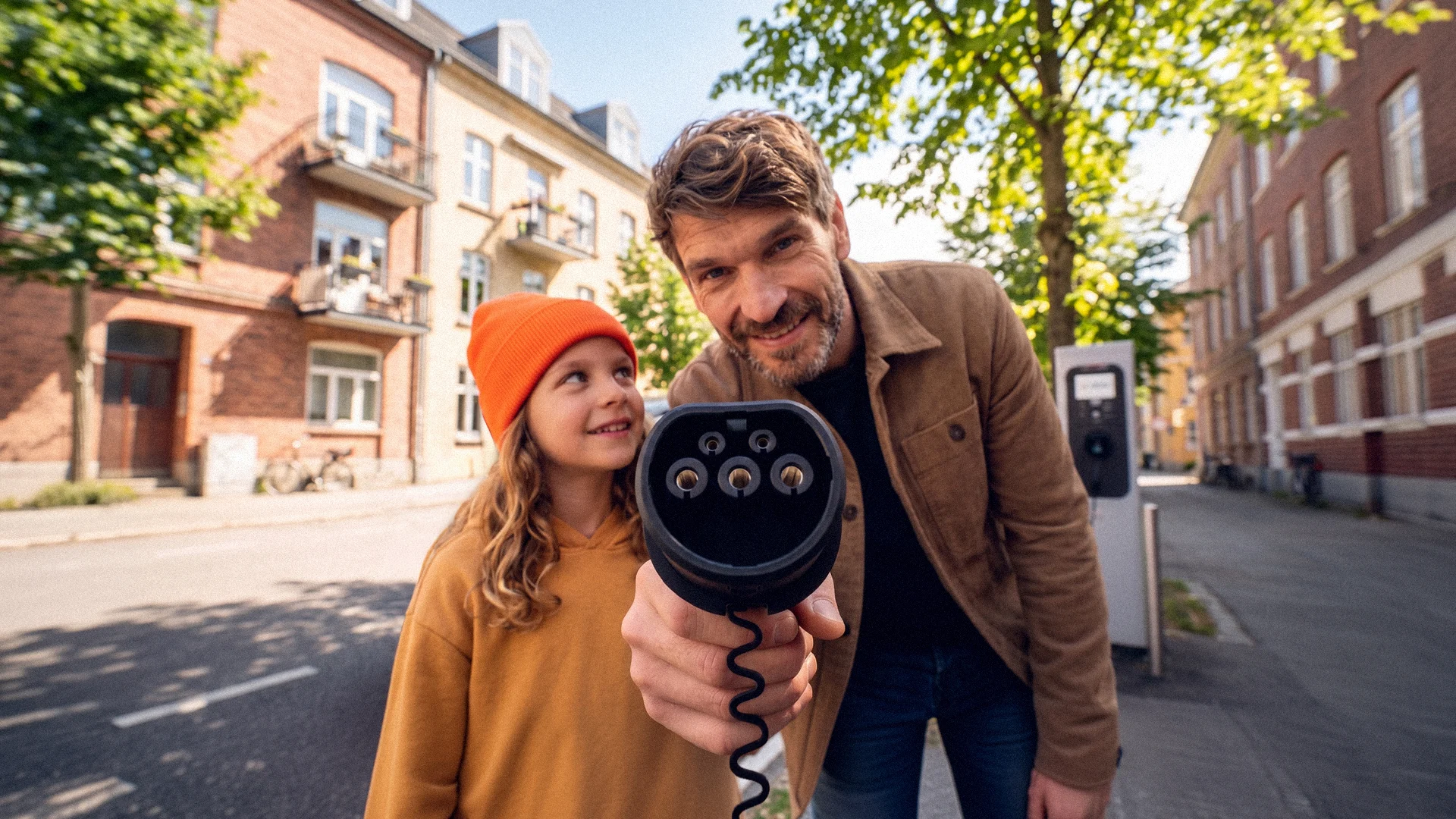 Ein Mann hält einen Typ-2-Ladestecker für ein Elektroauto direkt in die Kamera. Ein Kind im orangefarbenen Beanie schaut ihn an. Im Hintergrund ist eine Straße mit Backsteingebäuden und eine Ladesäule zu sehen. Das Bild trägt den Namen