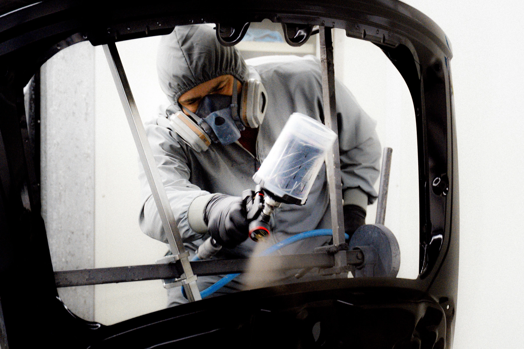 Ein Mitarbeiter des Autohauses steht vor einem Karosserie-Teil in einer Lackierkabine. Er hat einen Schutzanzug und eine Schutzmaske an. In seiner rechten Hand hält er eine Farb-Pistole, mit der er Farbe auf das Bauteil aufträgt.