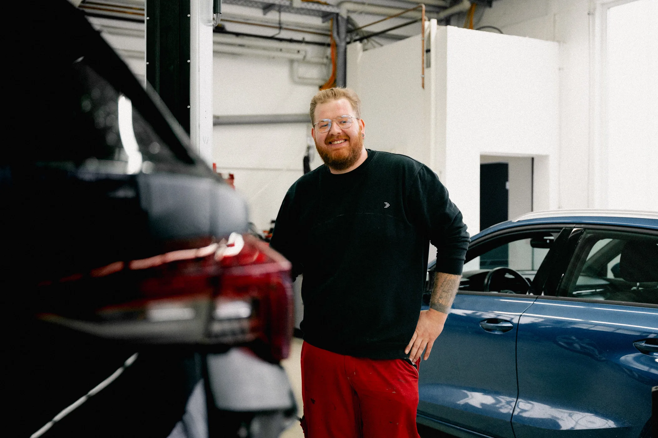 Ein Mitarbeiter des Autohauses steht zwischen zwei Autos in der Werkstatt. Er hat Arbeitskleidung an und lächelt geradeaus in die Kamera.