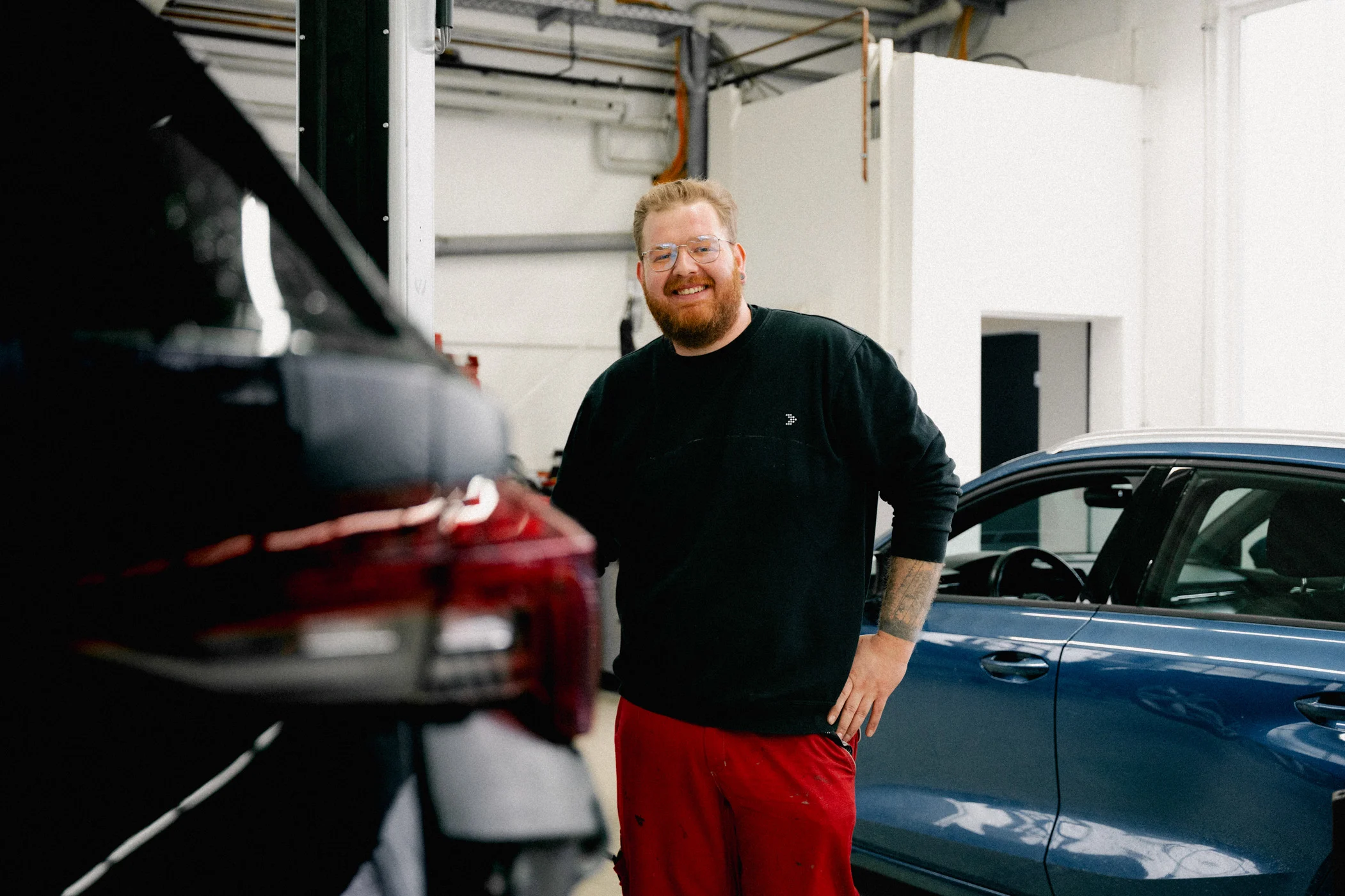 Ein Mitarbeiter des Autohauses steht zwischen zwei Autos in der Werkstatt. Er hat Arbeitskleidung an und lächelt geradeaus in die Kamera.