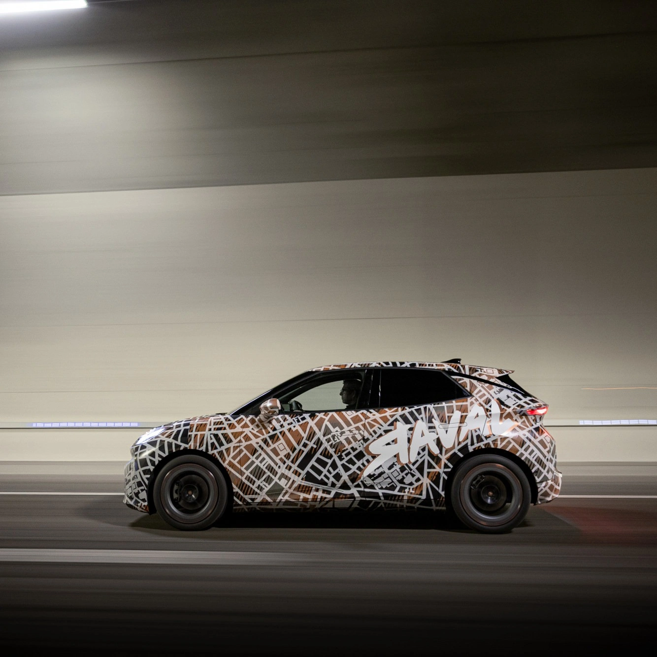 Seitenansicht eines CUPRA Raval Prototyps bei einer dynamischen Fahrt durch einen Tunnel. Das kompakte Elektroauto ist mit einer auffälligen schwarz-weiß-braunen Tarnfolie mit „RAVAL“-Schriftzug foliert, die das Design vor der Markteinführung verbirgt.