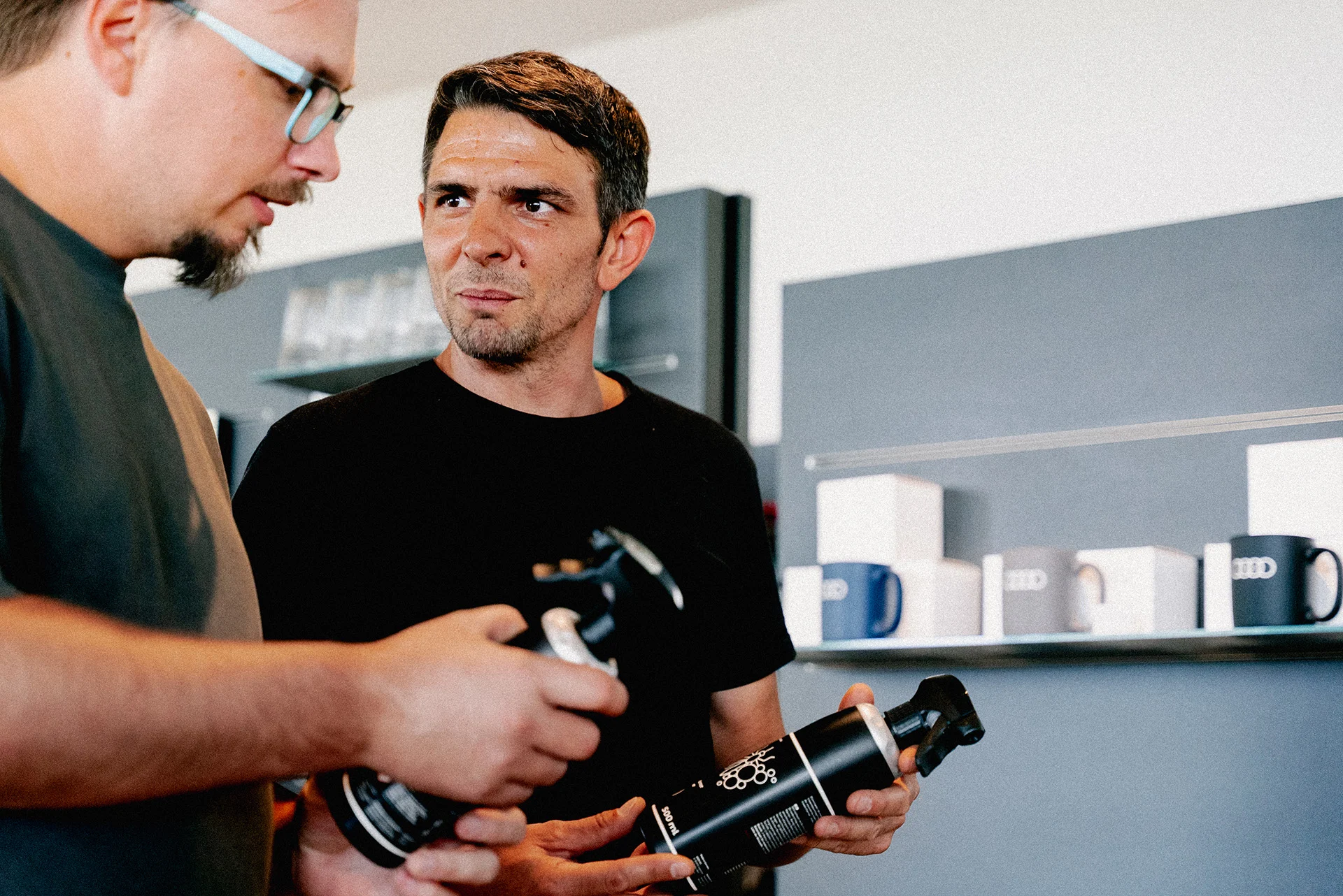 Ein Mann im schwarzen T-Shirt berät einen männlichen Kunden mit Brille, während beide jeweils eine schwarze Sprühflasche in der Hand halten. Im Hintergrund sind dunkle Regale mit Tassen zu erkennen.