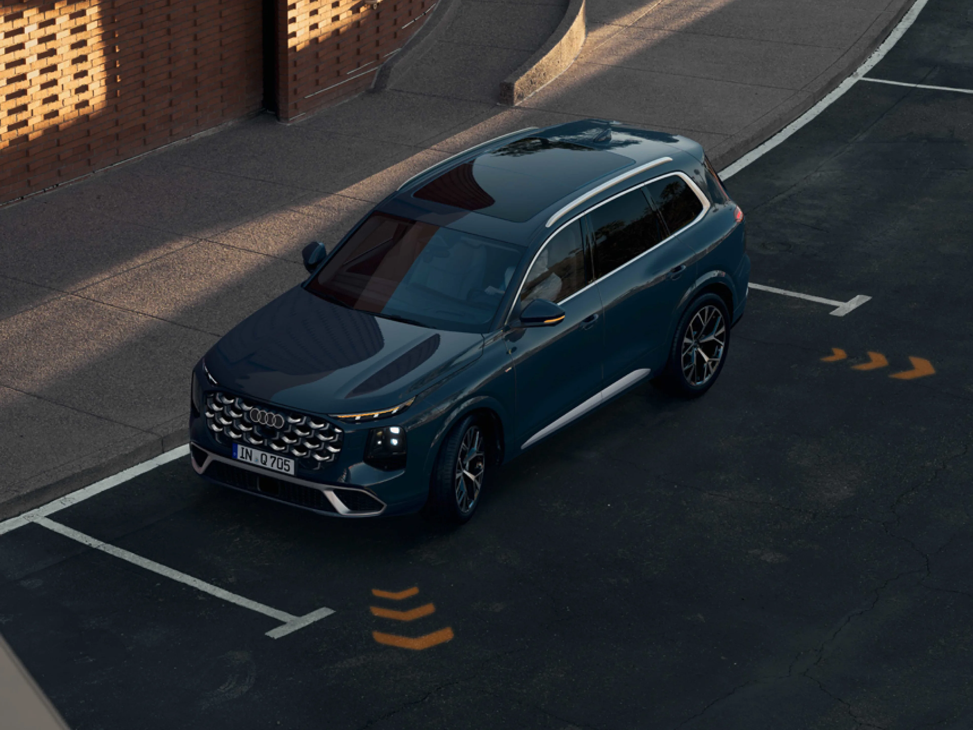 Ein graublauer Audi Q7 aus der Vogelperspektive auf einem Parkplatz im Abendlicht. Zu sehen sind das Panoramadach, der markante Kühlergrill mit Audi-Ringen, das Kennzeichen „IN Q 705“ und gelbe Pfeilmarkierungen auf dem Asphalt.