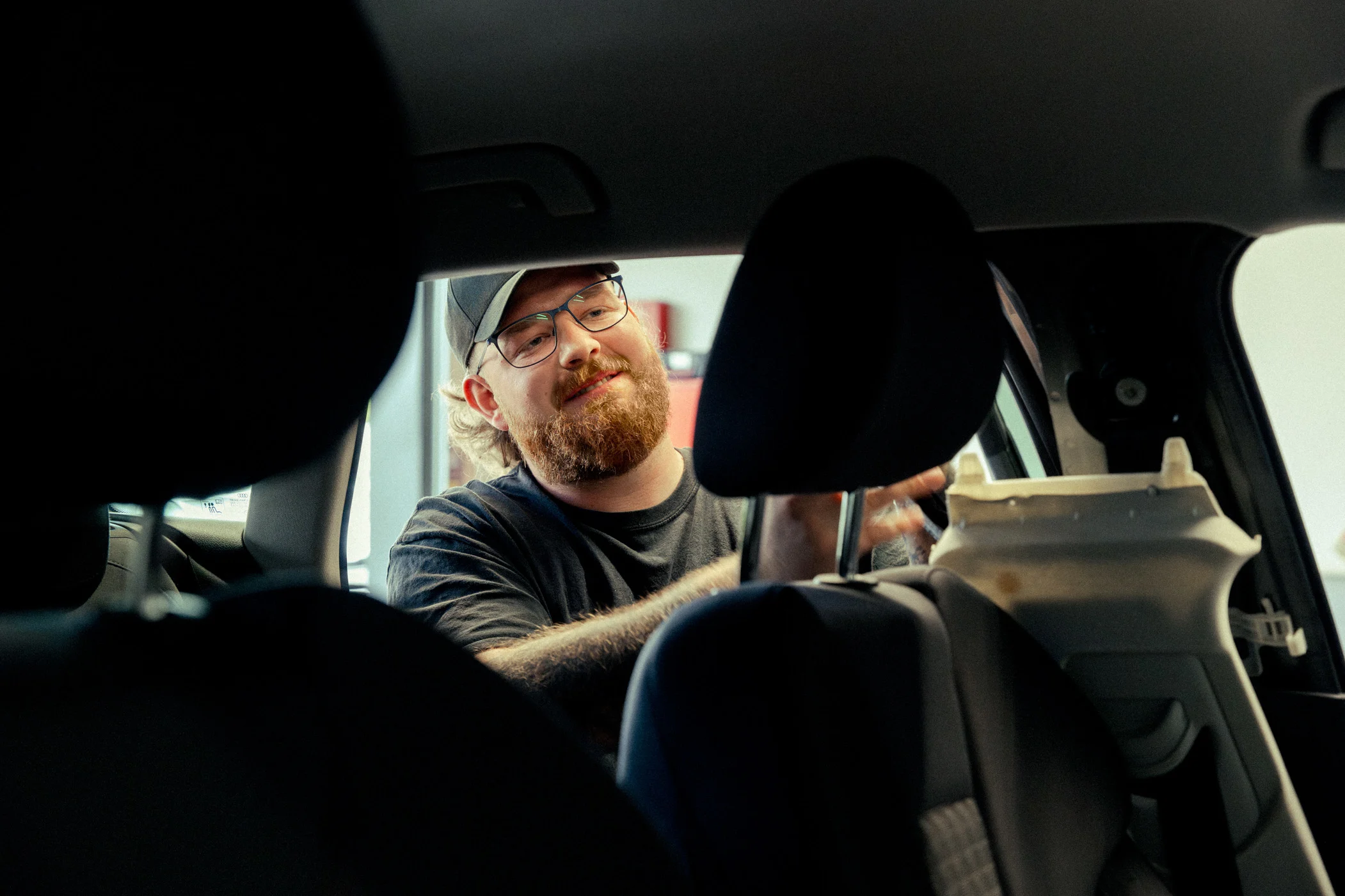 Werkstatt durchs Fahrzeug m Eine Aufnahme quer durch den Innenraum eines Fahrzeugs. An der offenen Tür des Fahrzeugs steht ein Werkstattmitarbeiter mit Brille und Basecap. Er hebelt ein Stück der Innenraumabdeckung an der B-Säule aus, um daran zu arbeiten.