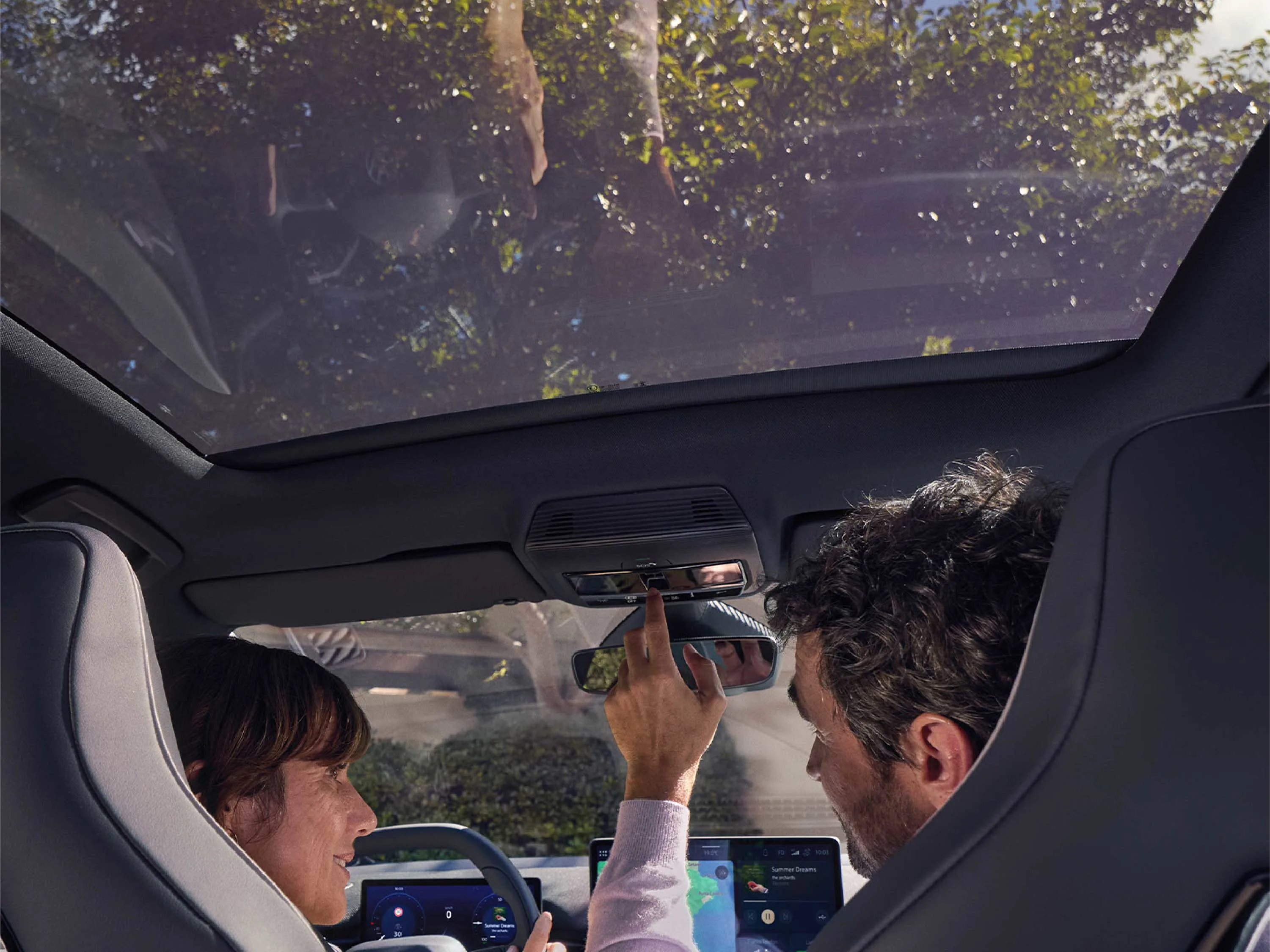 Blick von den Rücksitzen in den VW ID.3. Fahrer und Beifahrer sitzen unter einem großen Panorama-Glasdach, in dem sich grüne Bäume spiegeln. Ein Arm greift nach oben zur Dachbedieneinheit; im Hintergrund ist das Navigations-Display erkennbar.