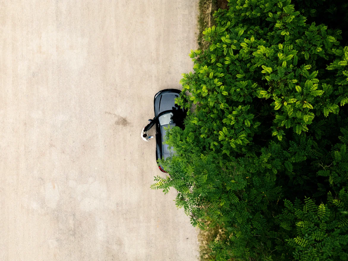 Fahrzeug Vogelperspektive Wald Eine Drohnenaufnahme eines schwarzen Audi RS e-tron GTs. Die Drohne schwebt genau über dem Auto und filmt senkrecht nach unten. Eine Person öffnet die Fahrertür des Autos. Ein Teil des Fahrzeugs und die rechte Seite des Bildes wird von Baumkronen verdeckt.