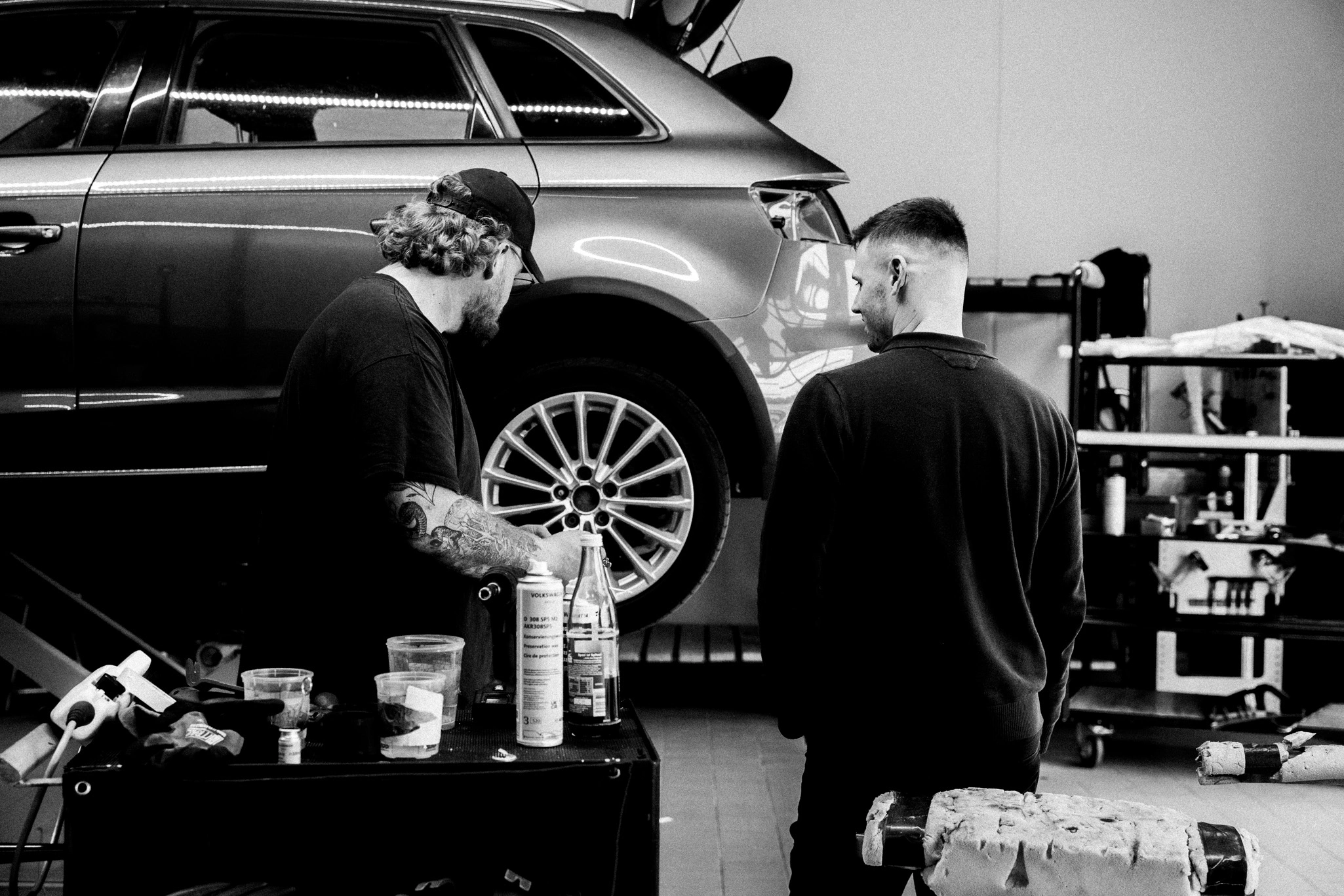 Man sieht die Seitenansicht eines Fahrzeuges, ab der B-Säule nach hinten, das auf einer Hebebühne steht. Davor stehen zweit Werkstatt-Mitarbeiter und ein Werkzeugkasten. Die Mitarbeiter bereiten Werkzeug vor, um am Fahrzeug zu arbeiten.