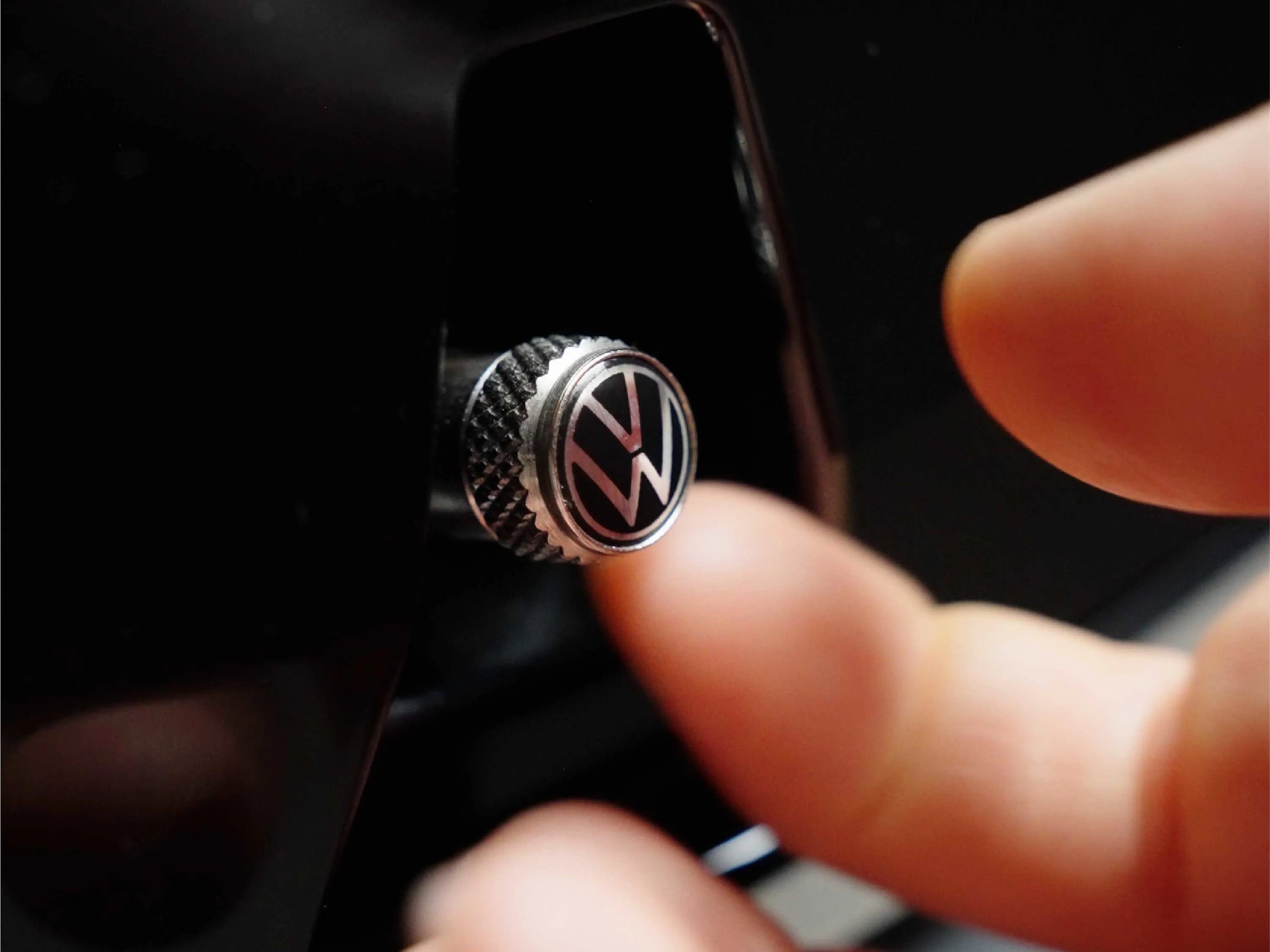 Nahaufnahme einer hochwertigen, silbernen Reifen-Ventilkappe mit eingeprägtem Volkswagen-Logo. Eine Hand im unscharfen Vordergrund greift nach dem Ventil, das an einer schwarzen Felge montiert ist.