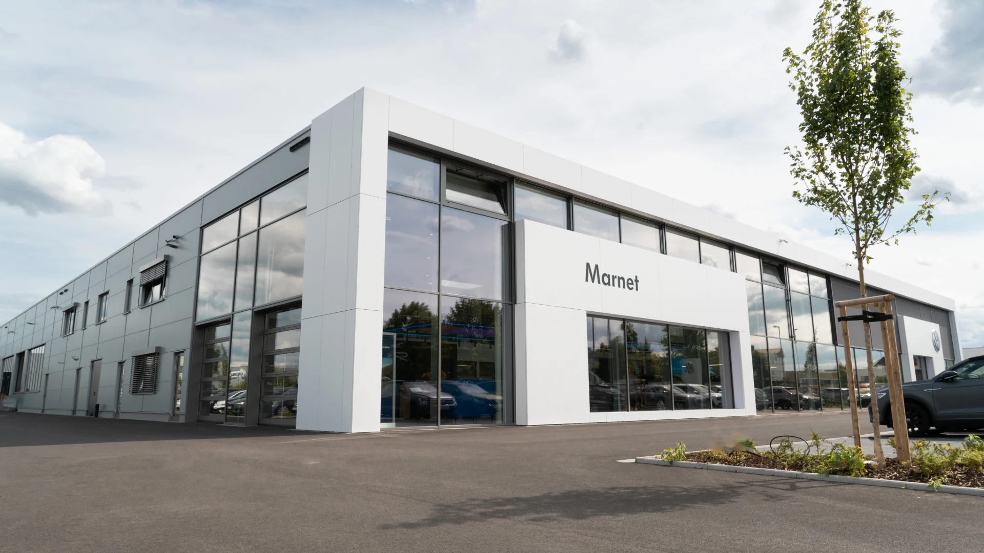 Volkswagen Wiesbaden Header Eine Aufnahme des Volkswagen Autohauses in Wiesbaden, dass das Gebäude und Volkswagen Fahrzeuge vor dem Gebäude, sowie die Einfahrt zeigt.