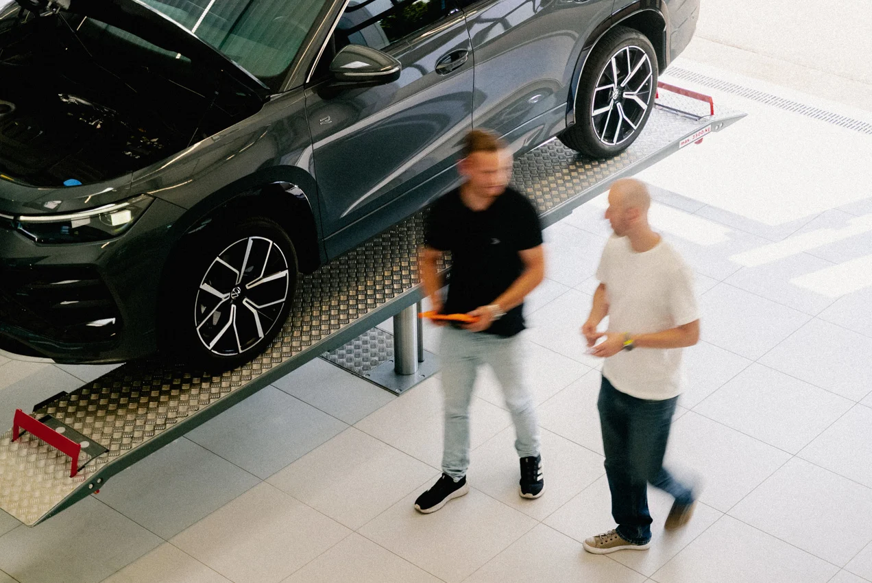 Zwei Männer laufen an einer Hebebühne in der Direktannahme des Autohauses vorbei. Die beiden unterhalten sich miteinander. Der Mann auf der linken Seite trägt ein AVEMO Polo-Shirt und hält ein Tablet zur Datenerfassung in den Händen.