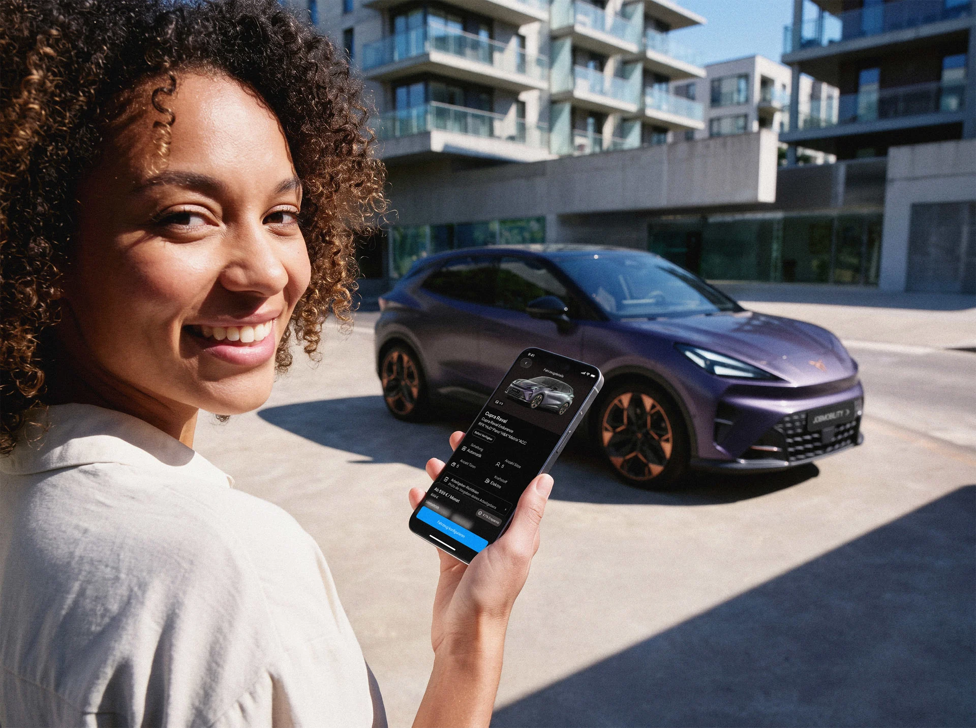 JobMobility – 7 Eine Frau zeigt ein Smartphone mit einer JobMobility-App-Anzeige für einen lila CUPRA Raval. Im Hintergrund steht derselbe CUPRA Raval mit dem Kennzeichen "JOBMOBILITY" vor städtischen Wohngebäuden.