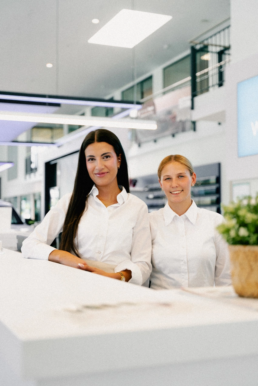 Eine Portrait-Aufnahme von zwei Service-Mitarbeiterinnen am Empfangsschalter des Autohauses. Beide lächeln direkt in die Kamera. Im Hintergrund sieht man noch einen Teil des Showrooms.
