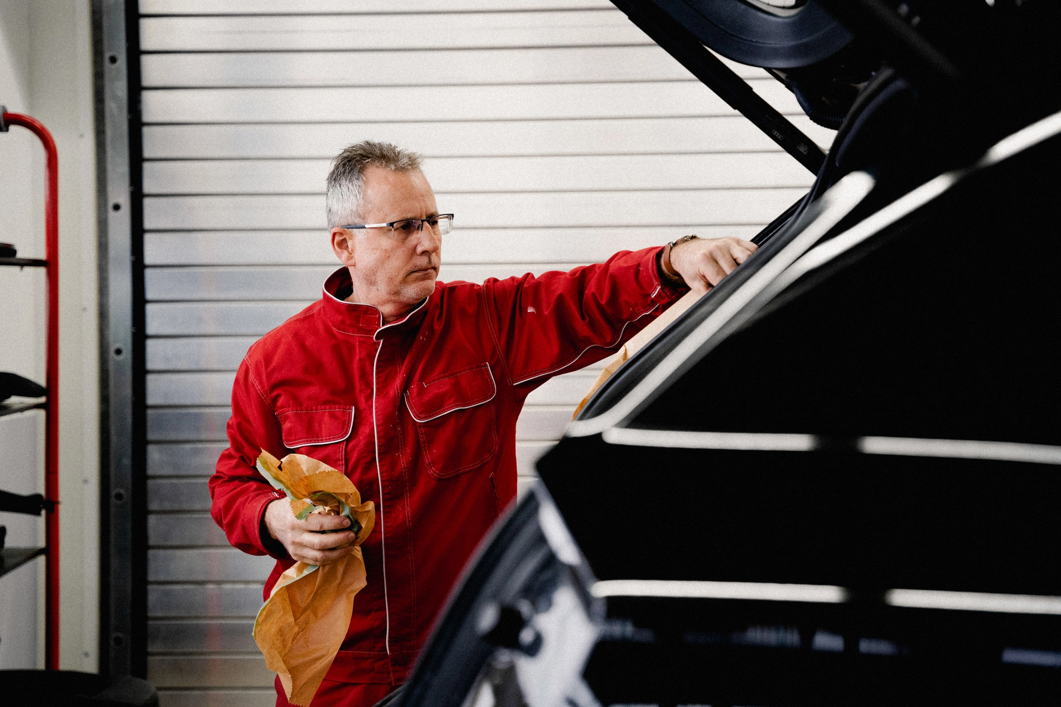 Zu sehen ist ein Werkstatt-Mitarbeiter in einem roten Overall. Er steht an der offenen Heckklappe eines Fahrzeuges und überprüft die Dichtungen des Kofferraums.