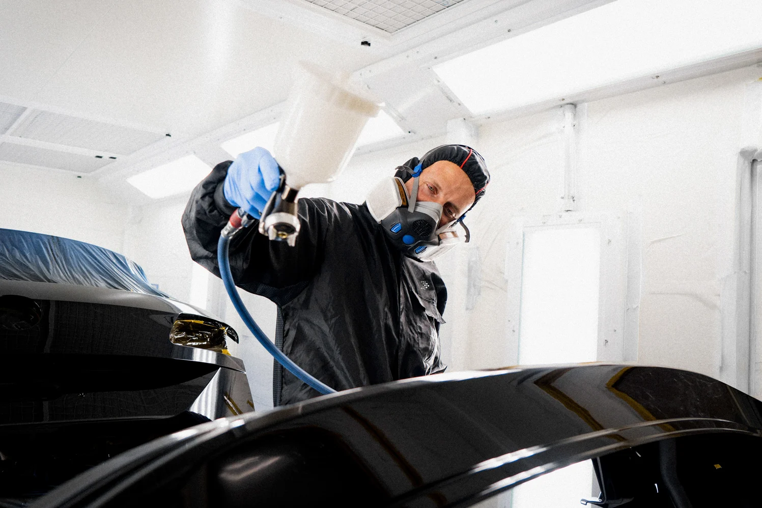 Ein Mitarbeiter des Autohauses steht vor einem Karosserie-Teil in einer Lackierkabine. Er hat einen Schutzanzug und eine Schutzmaske an. In seiner rechten Hand hält er eine Farb-Pistole, mit der er Farbe auf das Bauteil aufträgt.