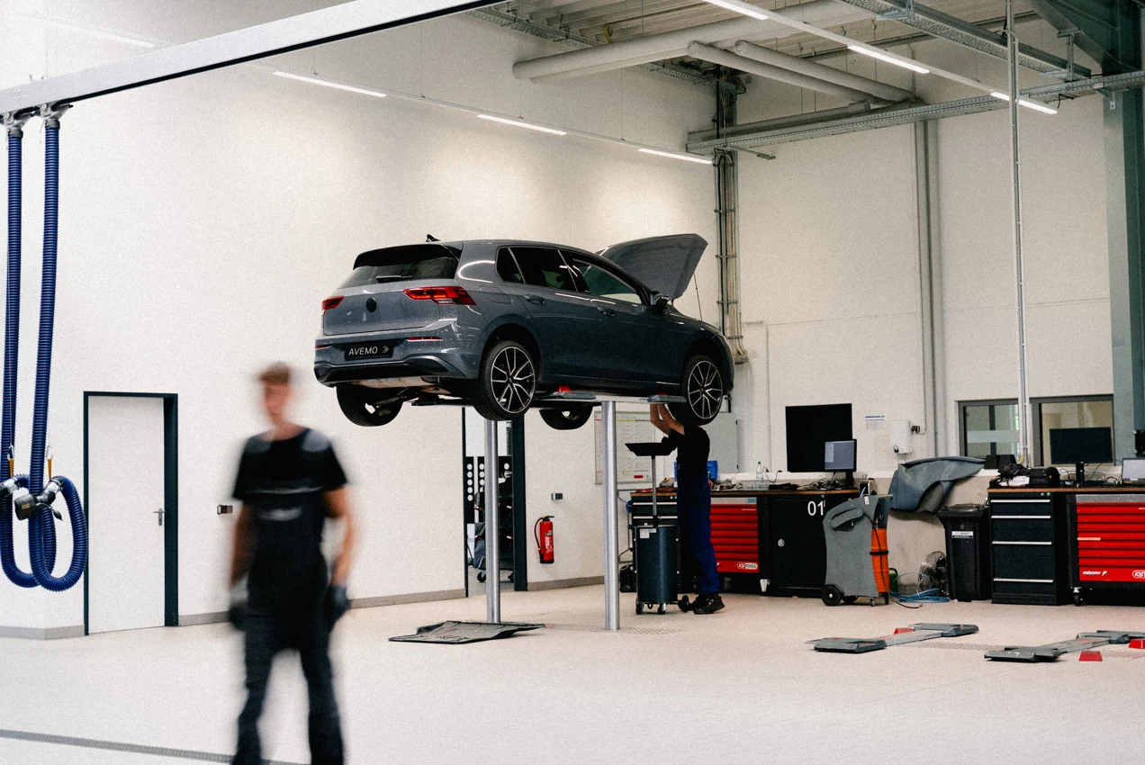 Zu sehen ist ein Auto auf einer Hebebühne des Autohauses in einer Werkstatt. Ein Werkstatt-Mitarbeiter steht unter dem Fahrzeug, um einen Ölwechsel durchzuführen. Ein anderer Mitarbeiter läuft im Vordergrund durch das Bild.