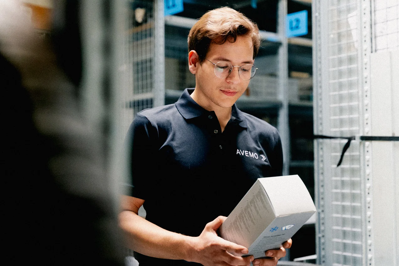 Ein Teiledienst-Mitarbeiter steht zwischen Regalen des Teiledienstes. Er trägt ein schwarzes AVEMO Polo-Shirt und eine Brille. Sein Blick ist auf ein Produkt in seinen Händen gerichtet, welches er zuvor aus dem Regal genommen hat.