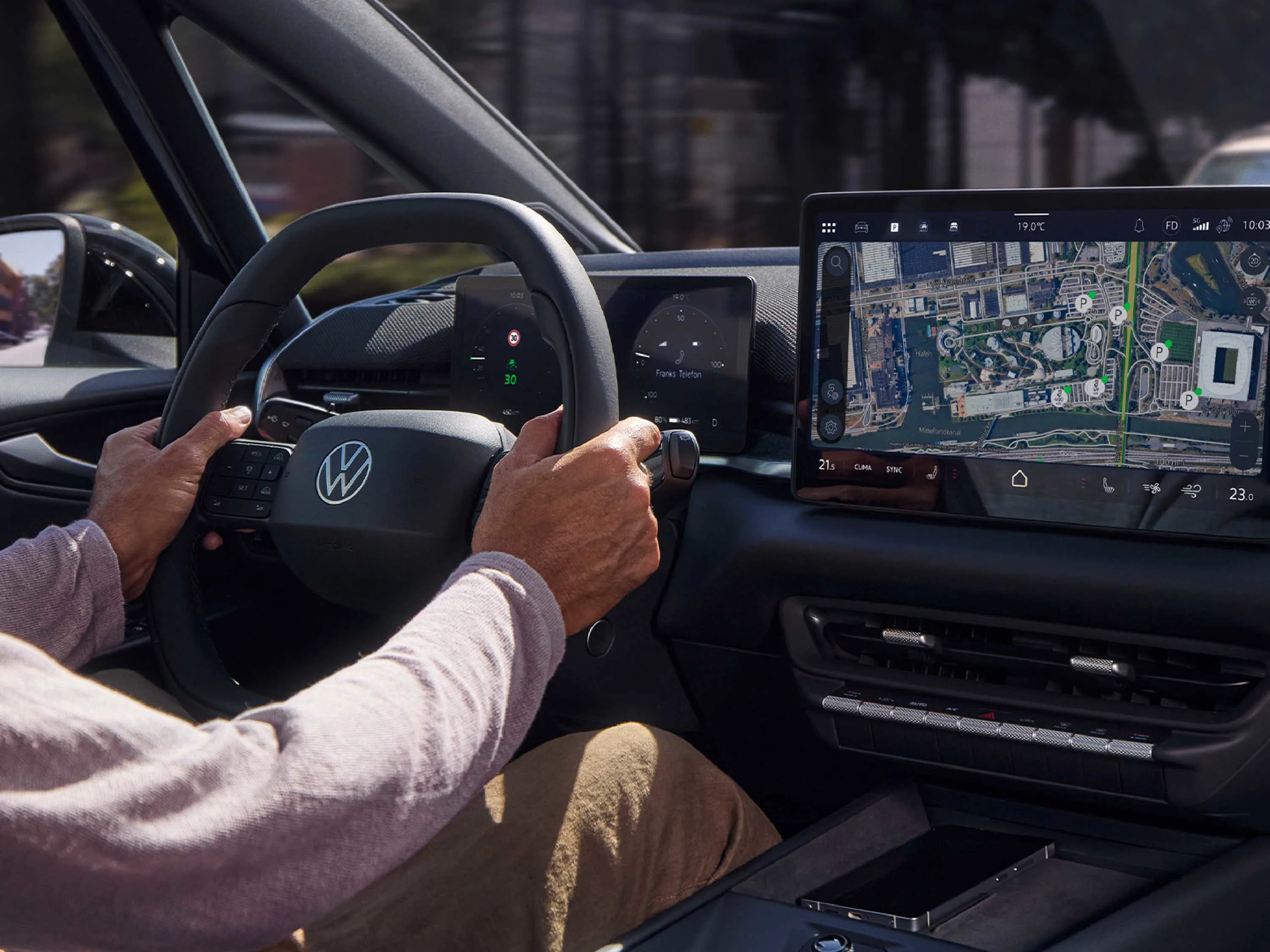 Perspektive vom Beifahrersitz auf das Cockpit eines VW ID.3 während der Fahrt. Ein Fahrer hat die Hände am Lenkrad; das große Display in der Mitte zeigt eine detaillierte Navigationskarte. Die Szene wirkt durch das Tageslicht hell und einladend.