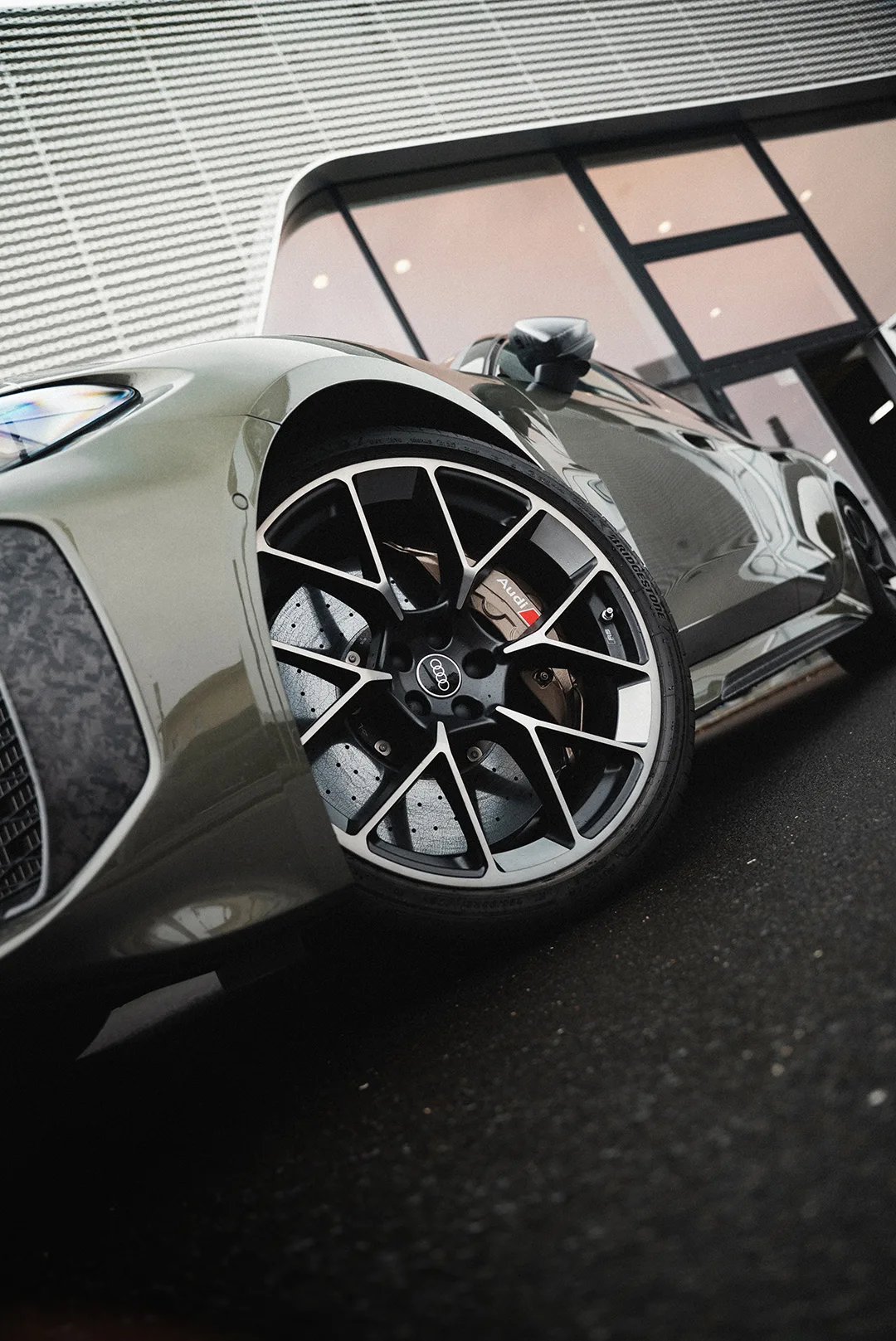Ein Foto eines grünen Audi Fahrzeugs mit starkem Fokus auf das vordere Rad der Fahrerseite. Im Hintergrund ist ein Audi Autohaus zu erkennen.