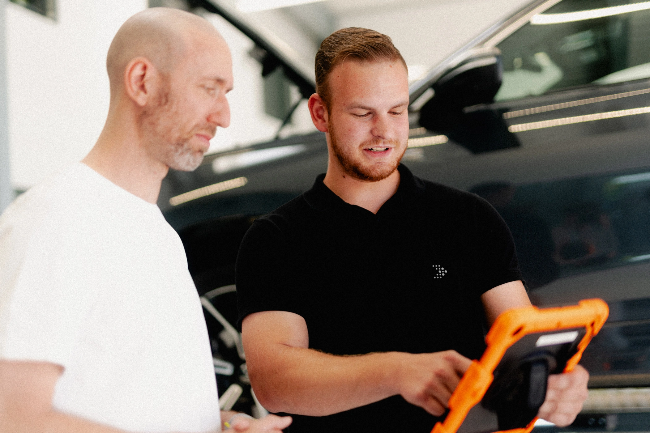 Direktannahme m, m Eine Aufnahme eines Serviceberaters in schwarzem AVEMO Poloshirt, der sich mit einem Kunden unterhält und auf das Display in seiner linken Hand deutet. Im Hintergrund befindet sich die Seite eines schwarzen Fahrzeuges.