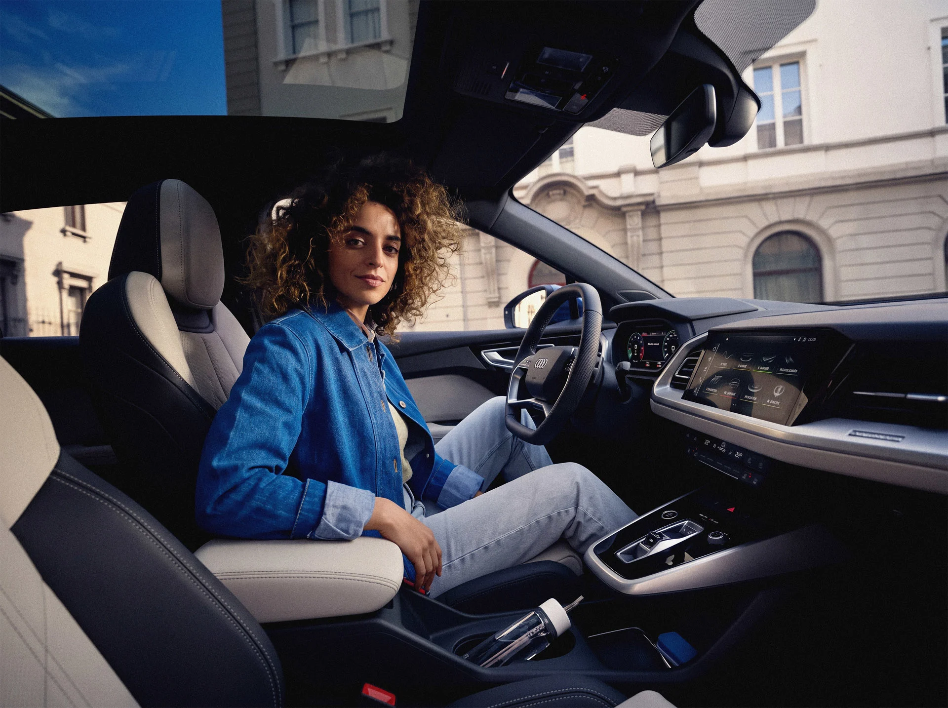 JobMobility - 2 Frau in Jeansjacke sitzt lässig im hochmodernen Cockpit eines Audi mit großem Infotainment-Display und Glasdach. Die Atmosphäre vermittelt den Komfort und die Freiheit der JobMobility App.
