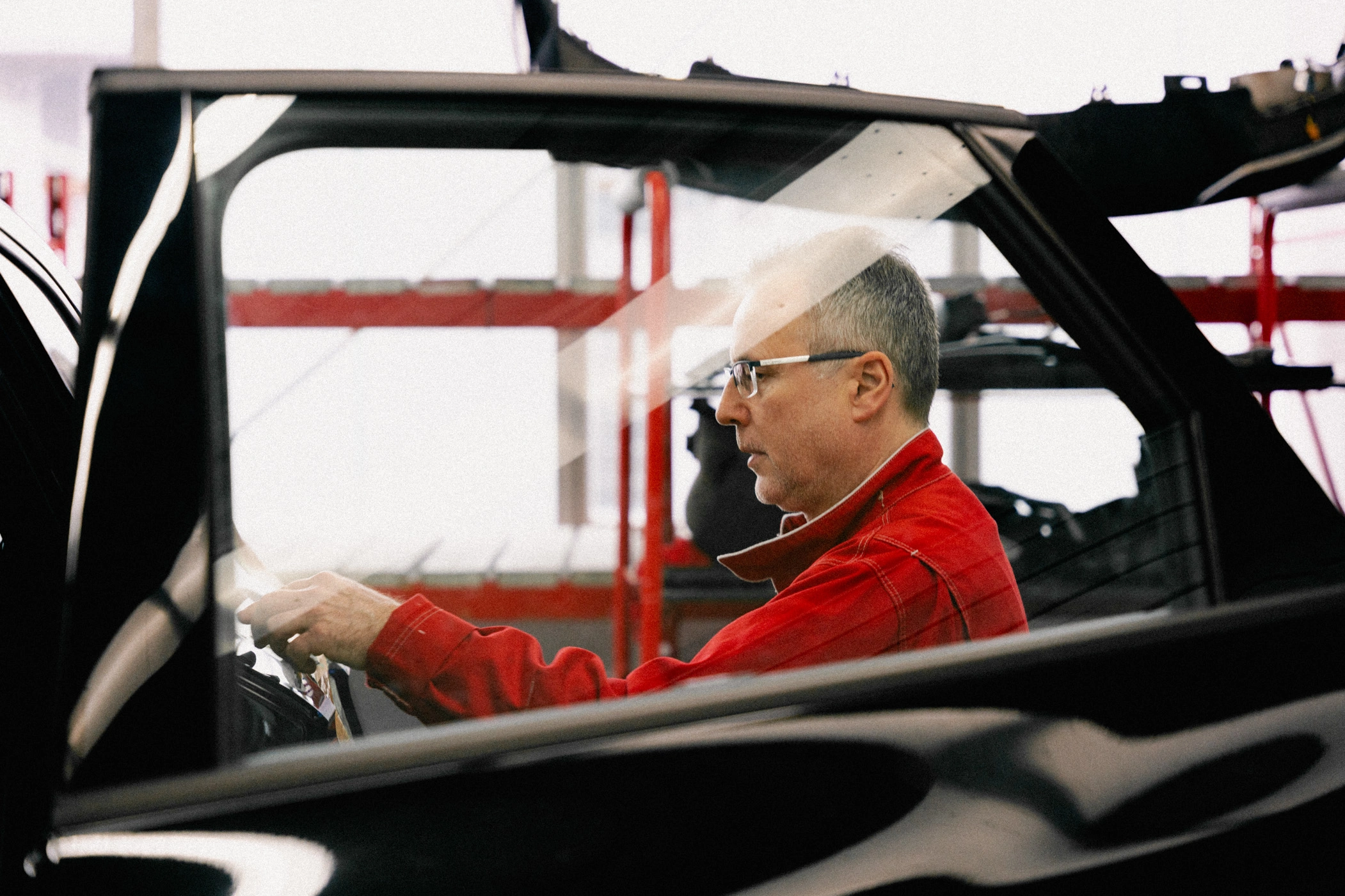 Eine Aufnahme durch die Fensterscheibe der offenen Fahrertür. Dahinter steht ein Werkstatt-Mitarbeiter seitlich am Fahrzeug und führt eine Prüfung des Lackes durch.