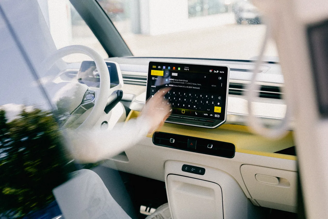 Eine Aufnahme eines Multimedia-Displays eines Neuwagens. Darauf zu sehen ist die Adresseingabe der Navigations-Funktion. Außerdem sieht man, wie sich die Hand des Fahrers in Richtung Display bewegt, um die Eingabe zu benutzen.