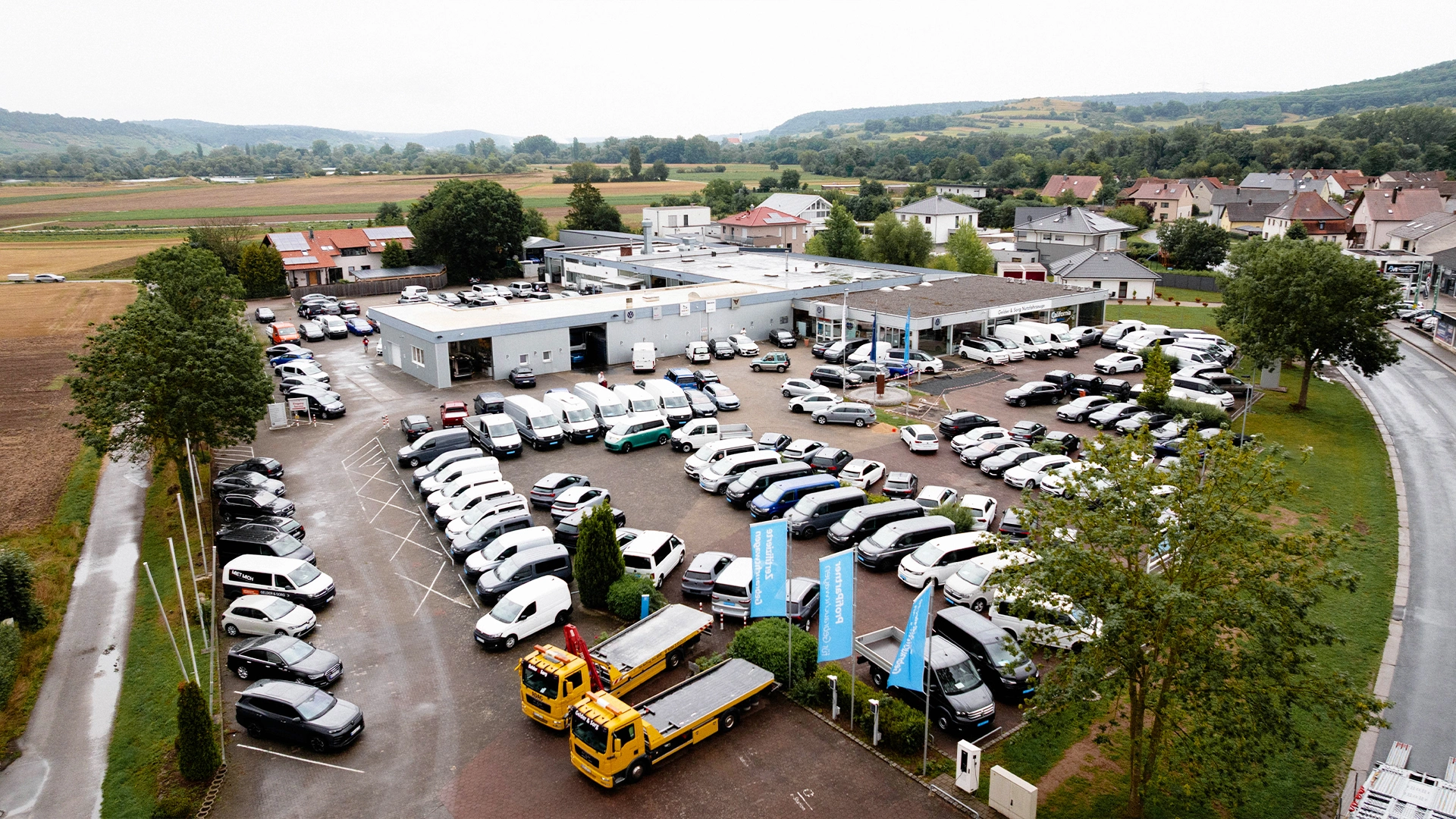Eine Aufnahme des Volkswagen Nutzfahrzeuge Autohauses in Sand am Main aus der Vogel-Perspektive, die das Gebäude, den großen Hof sowie Fahrzeuge vor dem Autohaus zeigt.