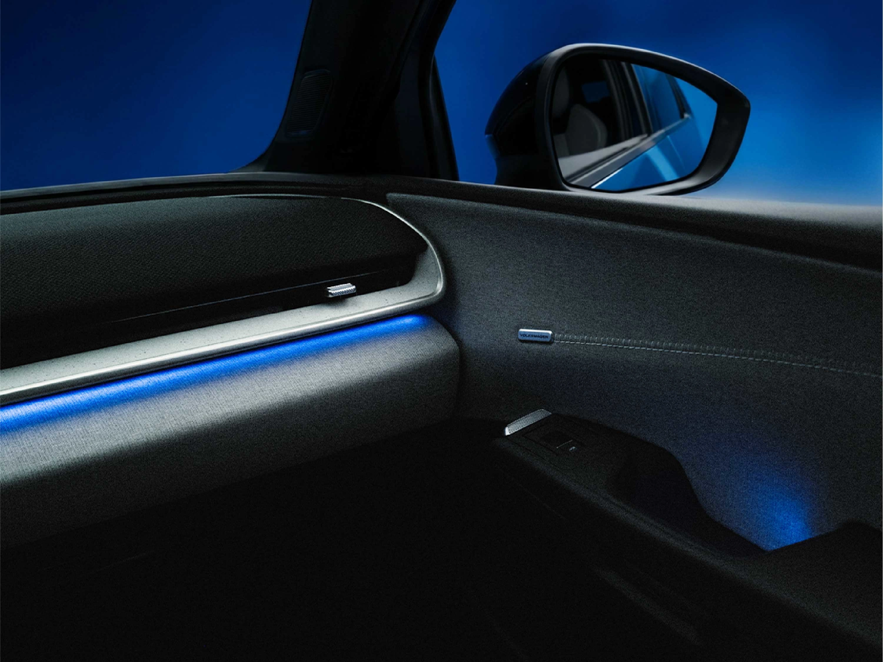 Detailaufnahme des Cockpits im VW ID.Polo mit Fokus auf die blaue Ambiente-Beleuchtung. Die Lichtleiste verläuft entlang des stoffbespannten Armaturenbretts und der Türverkleidung; im Hintergrund ist der Außenspiegel vor blauem Grund erkennbar.