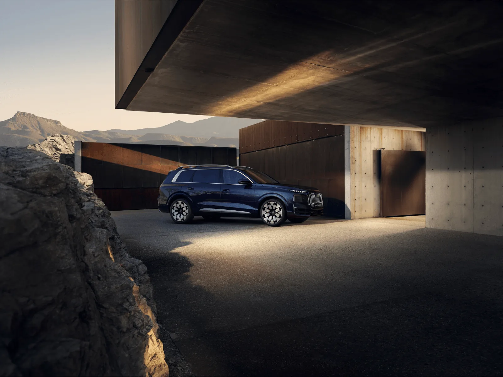 Seitenansicht eines dunkelblauen Audi Q9 vor moderner Betonarchitektur und einer Bergkulisse im Sonnenlicht.