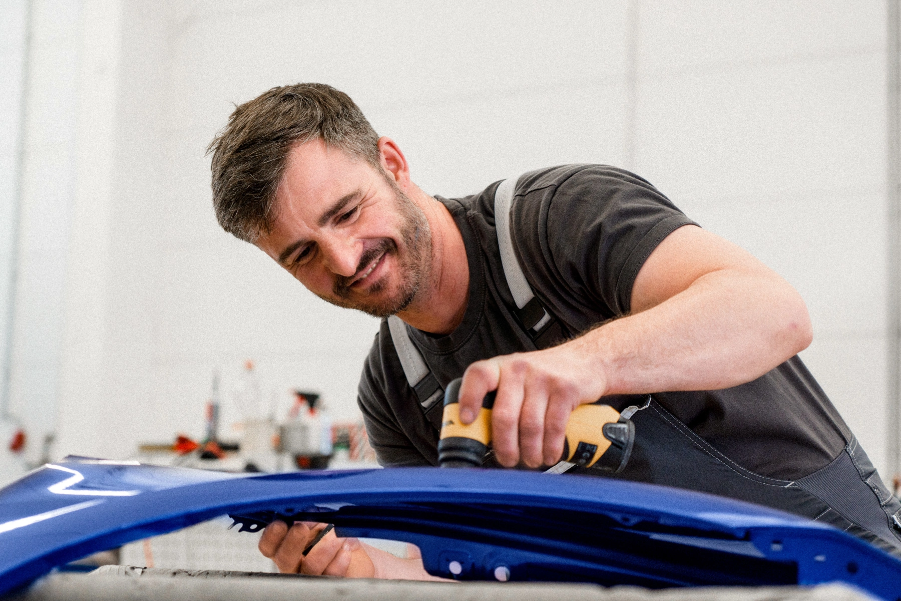 Zu sehen ist ein Werkstattmitarbeiter in einem grauen Overall. Er beugt sich über ein Karosserie-Teil, welches er mit der rechten Hand festhält und mit einem Poliergerät in der linken Hand poliert.