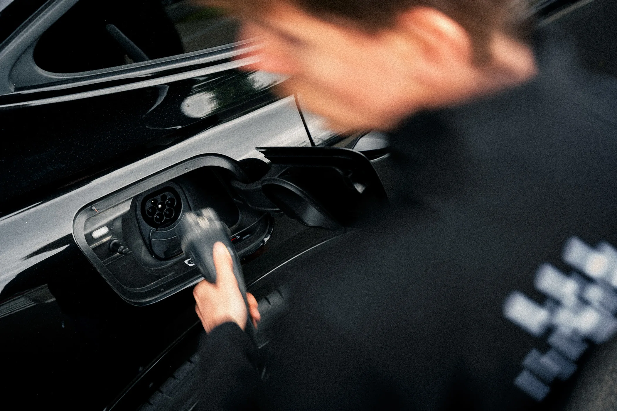 Eine Aufnahme einer offenen Ladebuchse eines E-Autos. Eine Person, die neben dem Auto steht, schließt gerade ein Ladekabel an, um den Ladevorgang zu starten.