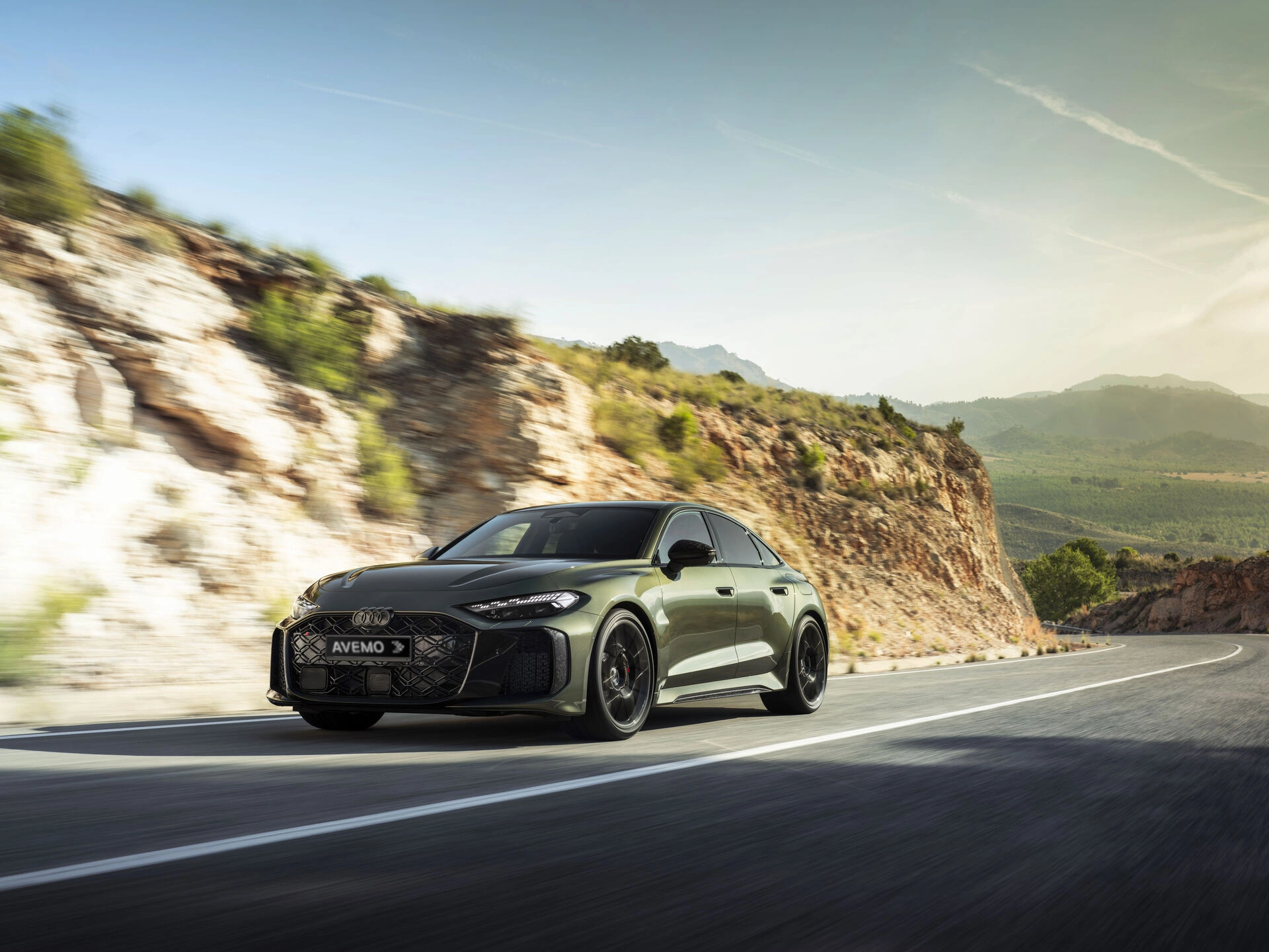 audi-rs-5-limousine-2026 Dynamische Frontansicht einer dunkelgrünen Audi RS 5 Limousine mit AVEMO-Kennzeichen während der Fahrt auf einer kurvigen Bergstraße. Das sportliche Fahrzeug mit markanten LED-Scheinwerfern ist vor einer sonnigen, felsigen Kulisse in Aktion zu sehen.