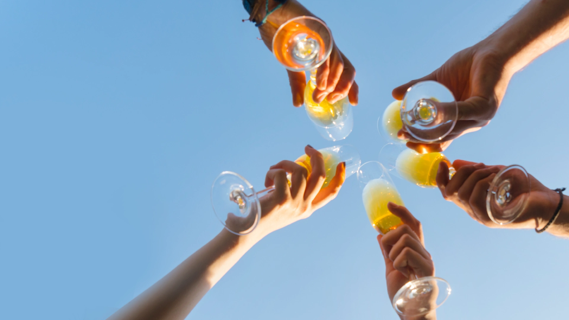 Fünf Personen stoßen mit Sektgläsern, die einen gelblichen oder orangen Inhalt haben, vor einem strahlend blauen Himmel an. Die Gläser treffen sich in der Mitte des Bildes.