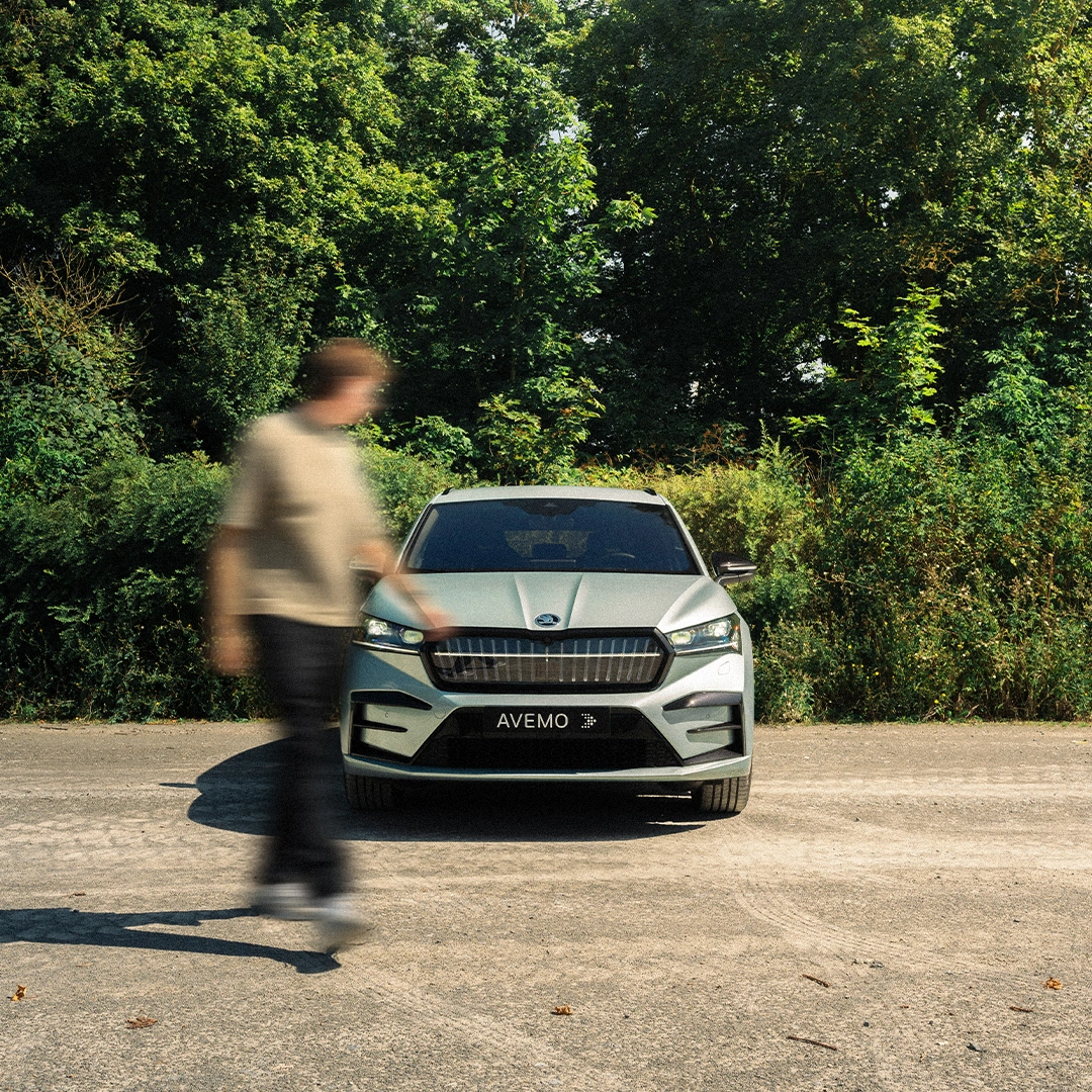 Fahrzeug Laufen Eine Aufnahme eines jungen Mannes, der an einem Škoda Fahrzeug vorbeiläuft. Das Fahrzeug steht vor einem grünen Stück Wald auf einem Schotterplatz.