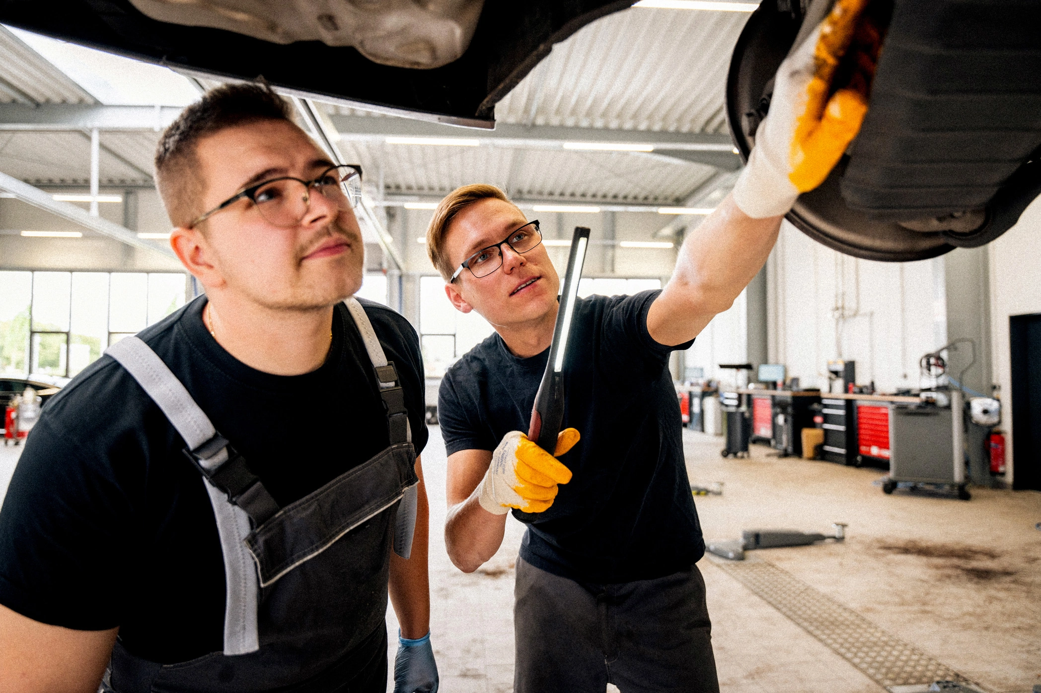 Werkstatt unterm Fahrzeug m,m Zwei Männer in Arbeitskleidung, die den Unterboden eines Autos auf einer Hebebühne begutachten. Einer der beiden Männer hält eine Werkstattlampe in der Hand und deutet auf ein Bauteil am Fahrzeug.