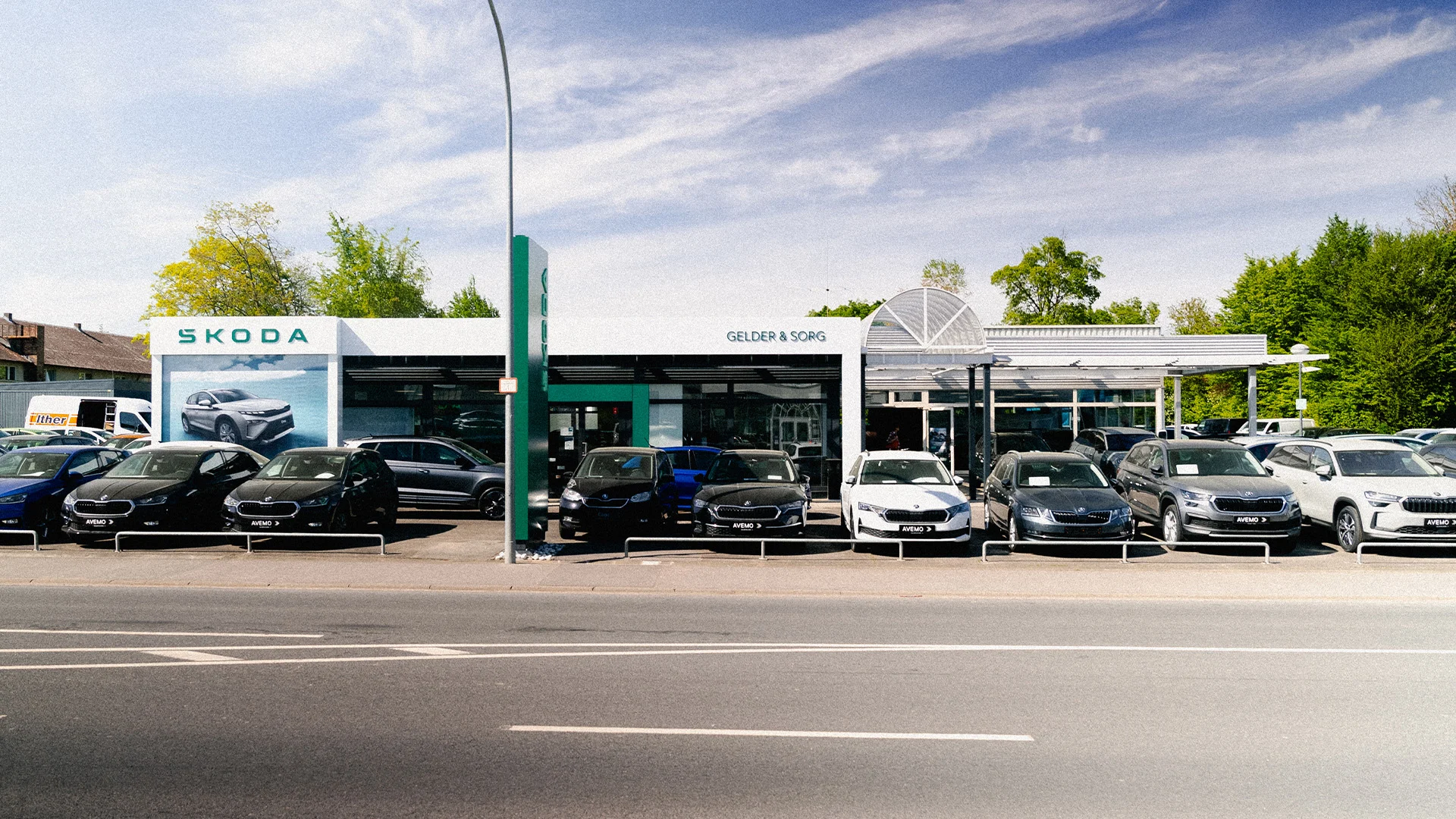 Eine Aufnahme des Škoda und Volkswagen Gebrauchtwagen Autohauses in Haßfurt, die das Gebäude und Škoda Fahrzeuge vor dem Gebäude, sowie die Einfahrt und die Zeiler Straße zeigt.